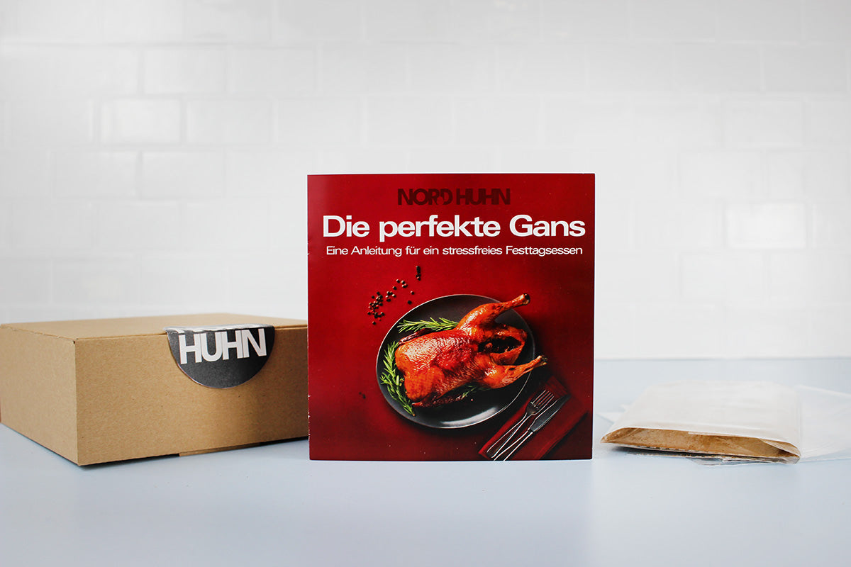 Rotes Rezeptheft mit Titel 'Die perfekte Gans' und Bild eines gebratenen Gänsebratens auf schwarzem Teller, daneben braune Pappschachtel mit Aufkleber 'HUHN' und braunes Papiertütchen