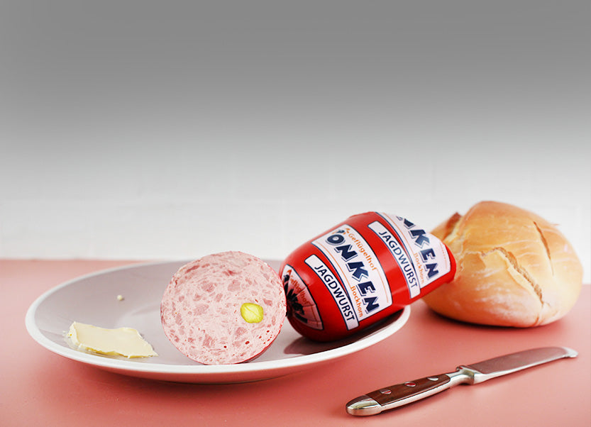 Aufgeschnittene Jagdwurst mit Pistazie auf weißem Teller, daneben Butter und Brötchen auf rosa Untergrund