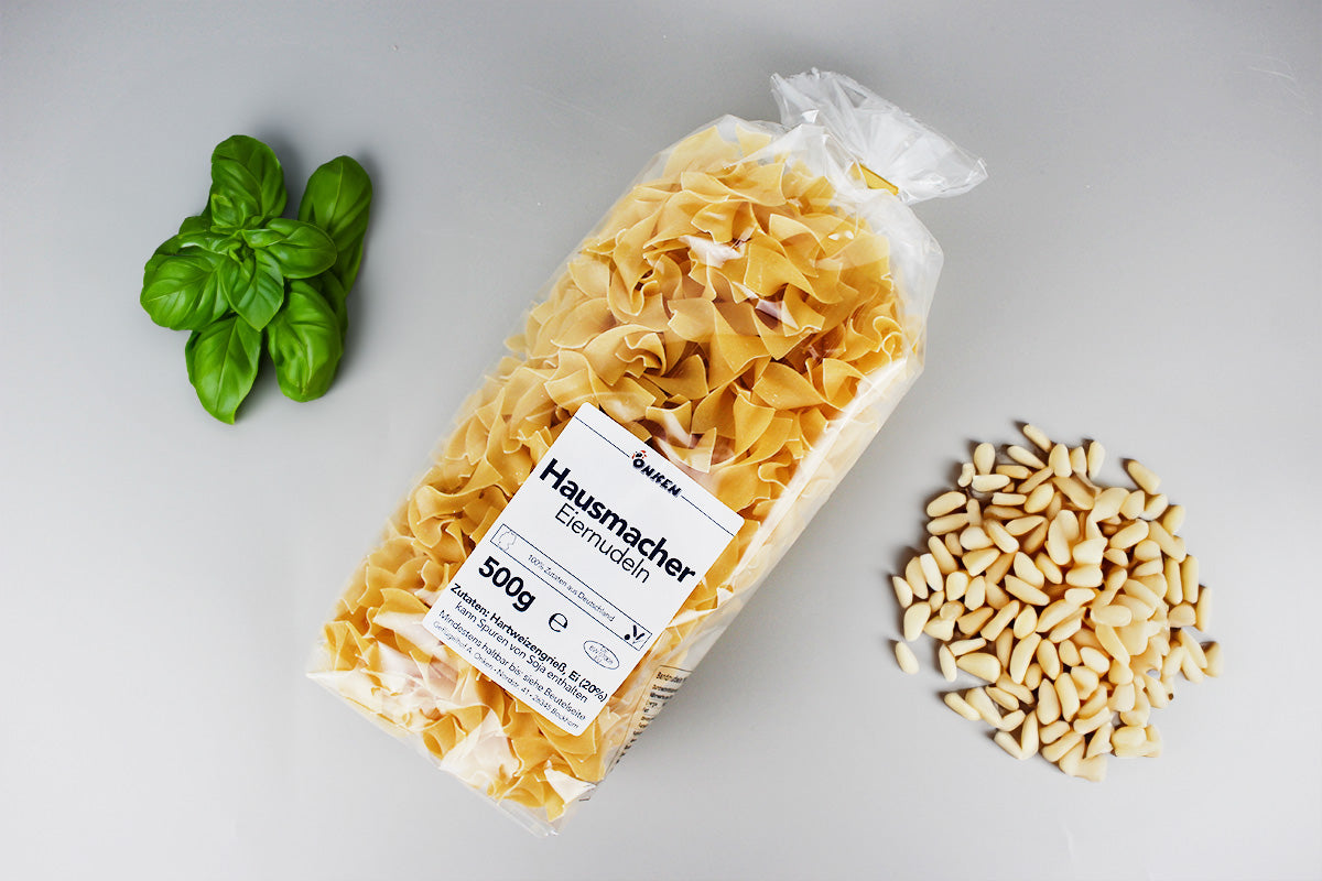 Packung mit Bandnudeln auf hellem Untergrund neben Pinienkernen und Basilikumblättern