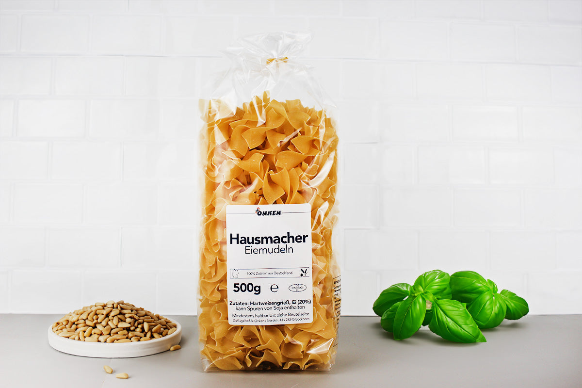Transparente Verpackung mit hausgemachten Eiernudeln neben einem Teller mit Kernen und einem Basilikumzweig