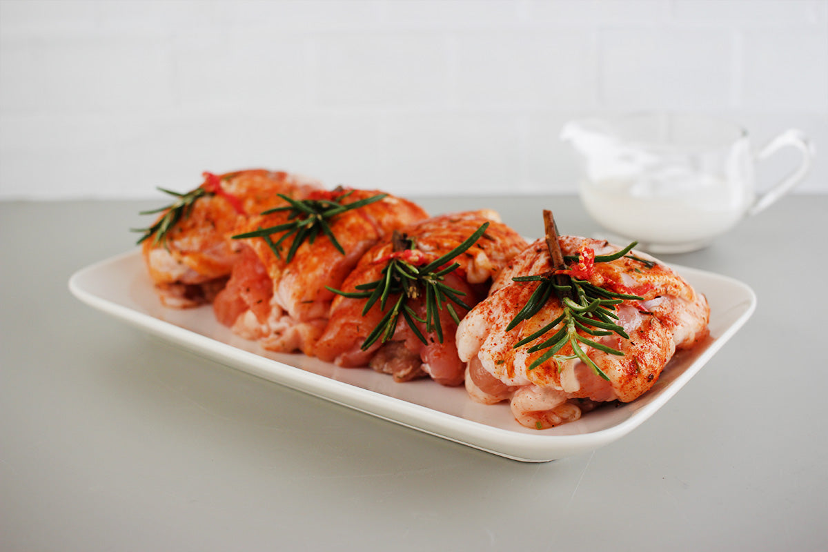 Vier gewürzte Hähnchenrollen mit Rosmarin zusammengebunden auf einer weißen Platte, begleitet von einem Glas frischem Rosmarin auf einer grauen Oberfläche vor einem weissen Fliesenspiegel