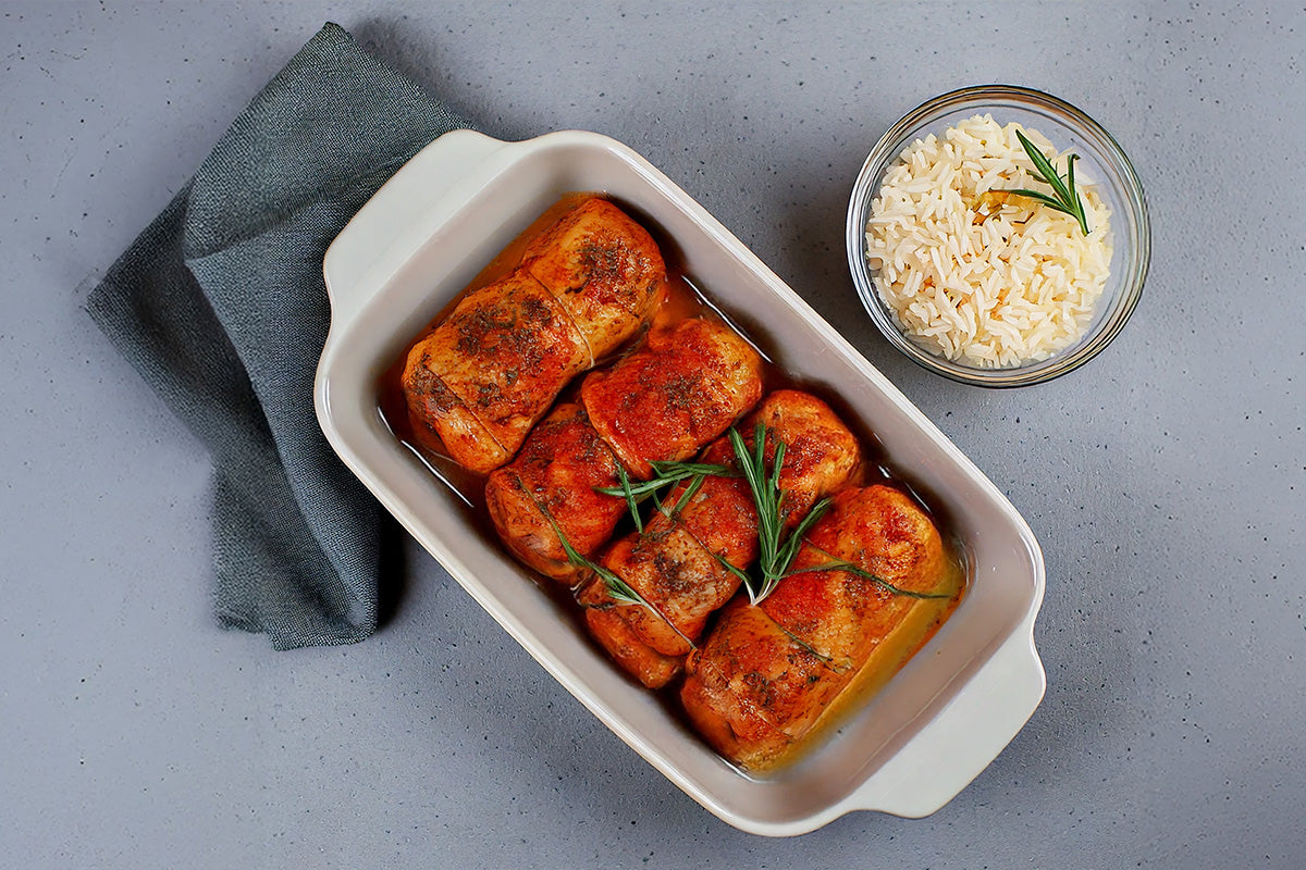 Vier gebackene Hänchenrollen mit Kräutern in einer weißen Schale, dazu eine Schüssel lockeren weißen Reis auf grauem Untergrund.