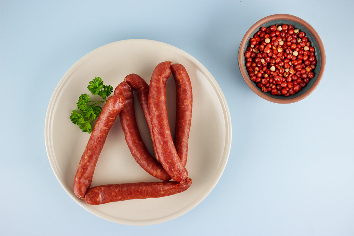 Sechas geräucherte Würste auf einem weißen Teller mit Petersilienzweig, daneben eine Schale mit roten Pfefferkörnern auf blauem Hintergrund