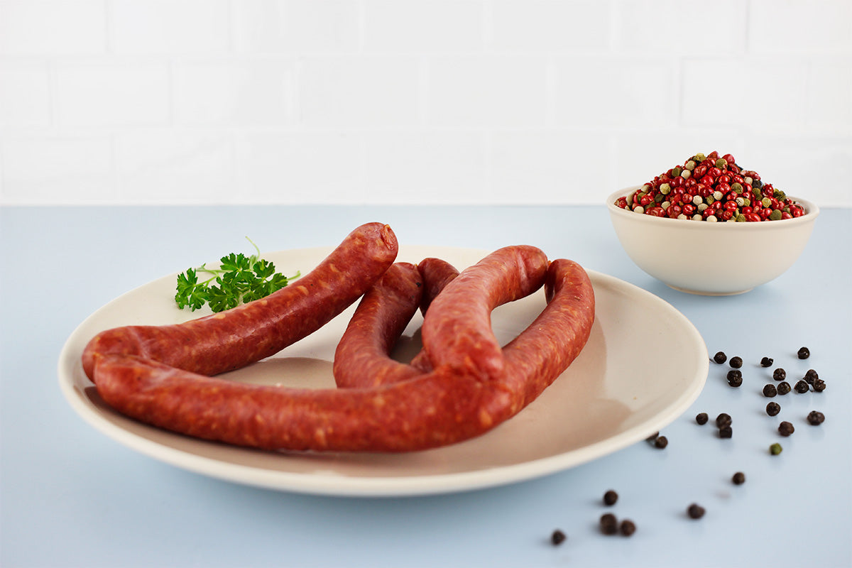 Fünf rote Bierknacker-Würste auf weißem Teller mit Petersilienzweig, daneben Schale mit buntem Pfeffer und verstreute schwarze Pfefferkörner