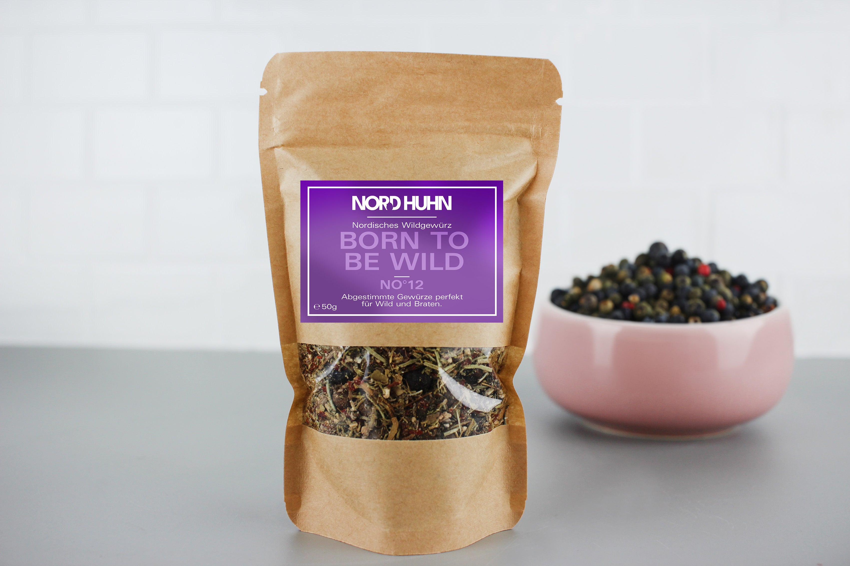 Braune Verpackung mit nordischem Wildgewürz 'Born to be Wild' von Nordhuhn, 50g, daneben rosa Schale mit bunten Pfefferkörnern