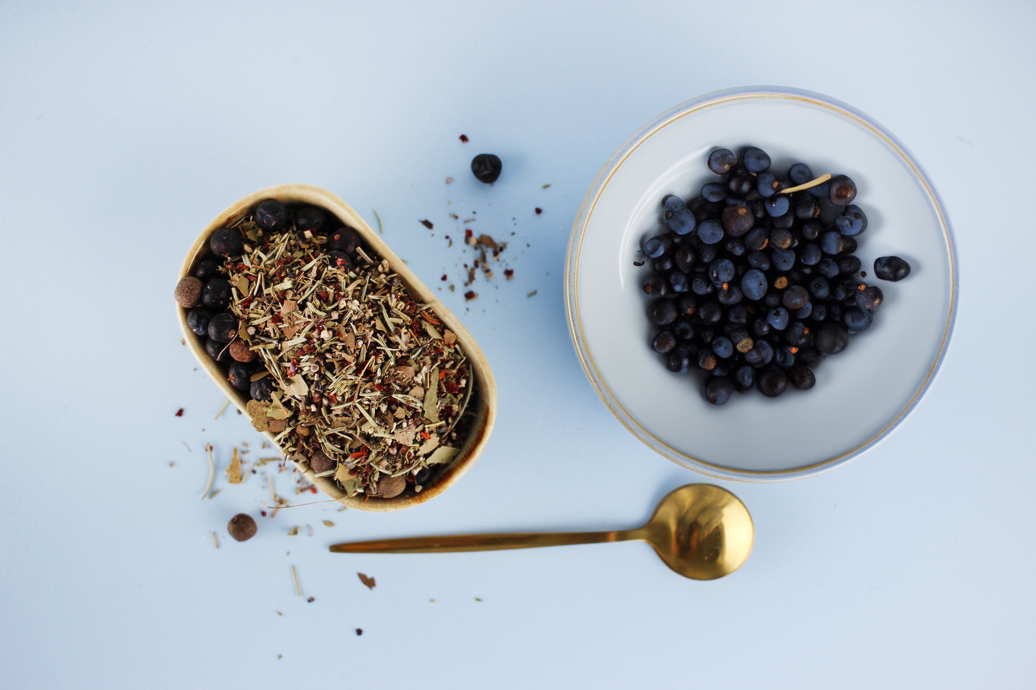 Schale mit getrockneten Gewürzen und Schale mit frischen schwarzen Beeren, daneben ein goldener Löffel auf hellem Hintergrund