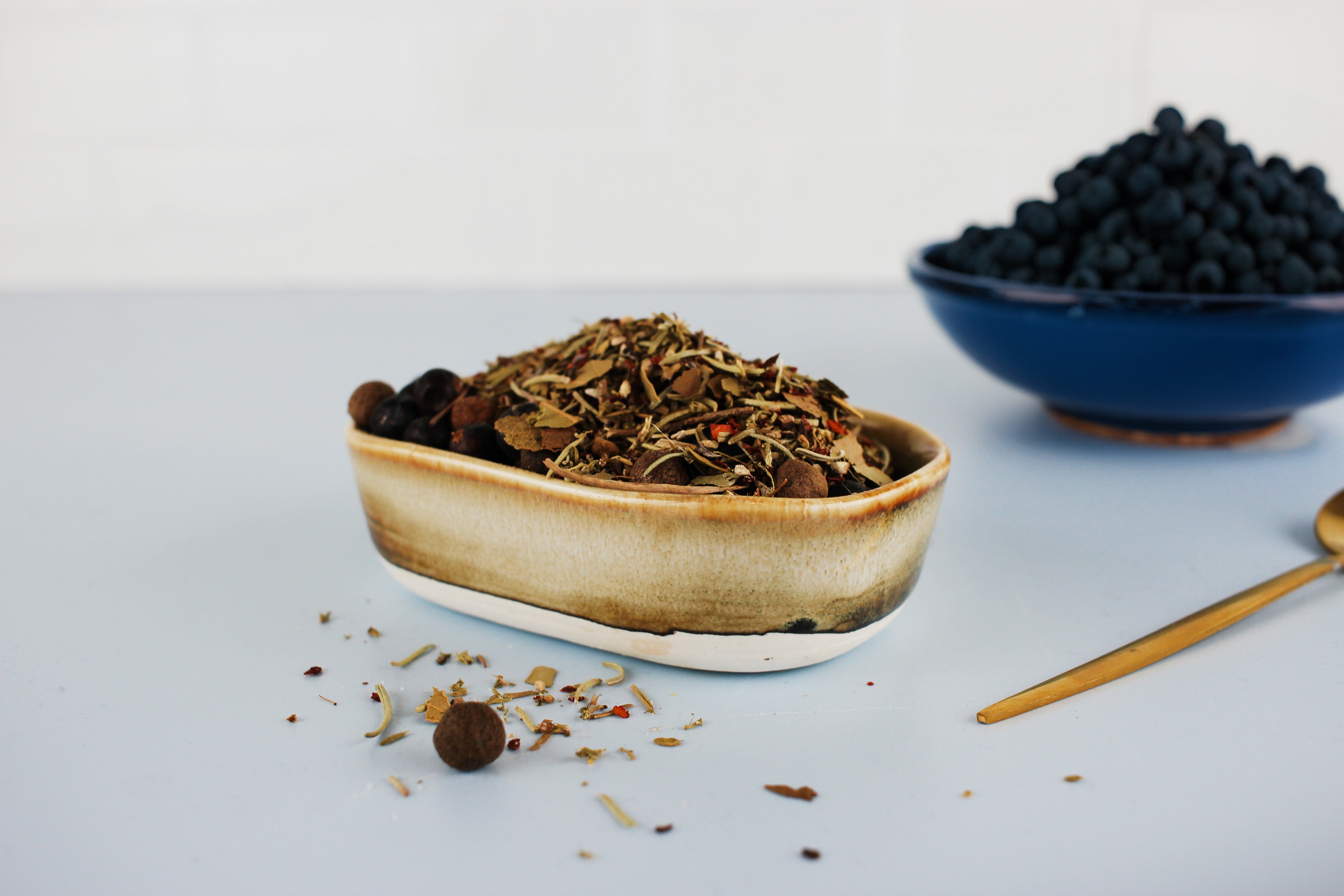 Schale mit gemischten Wildgewürzen und getrockneten Kräutern, daneben goldfarbener Löffel und blaue Schale mit schwarzen Beeren im Hintergrund