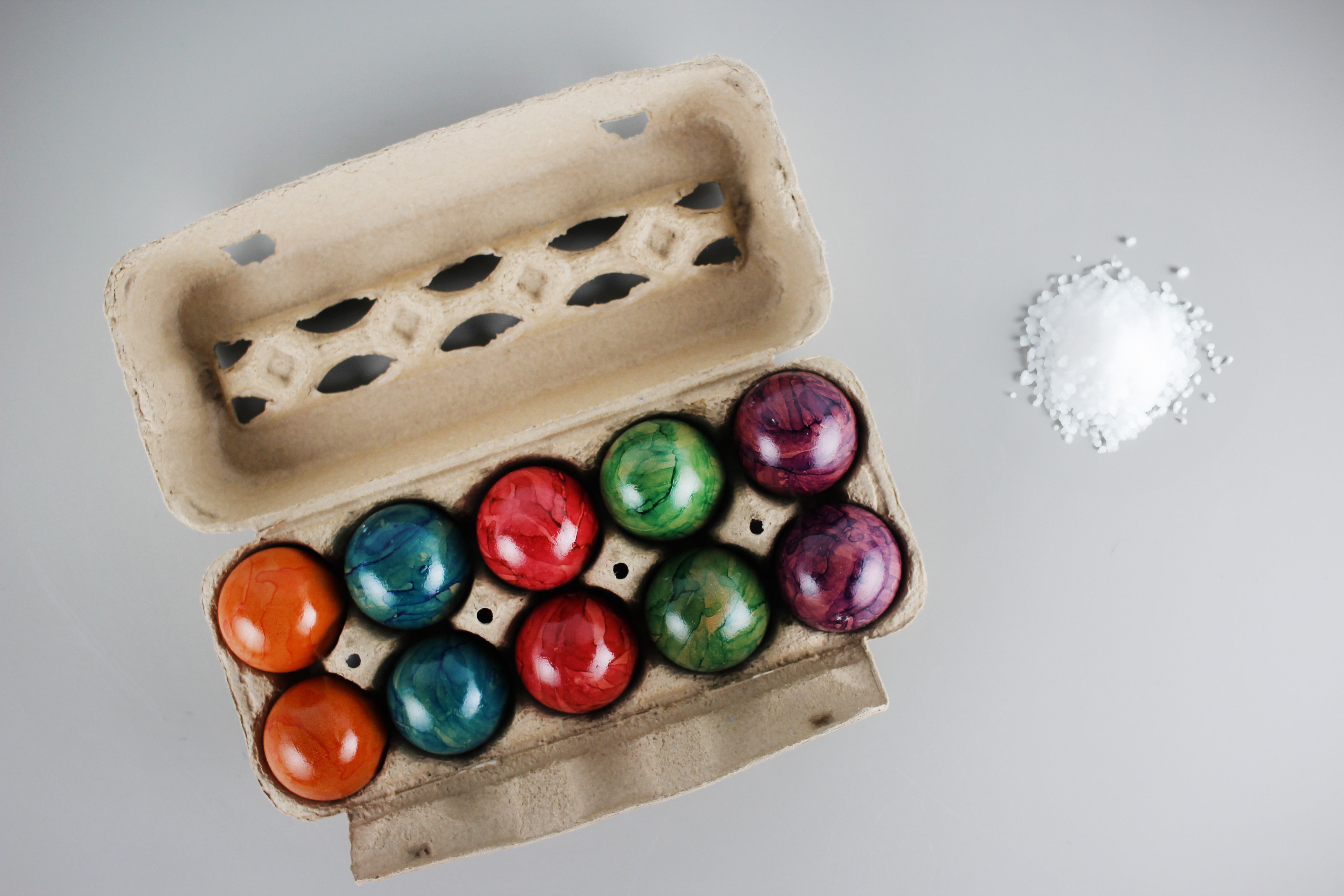 Ein Karton mit zehn bunten Ostereiern, daneben auf grauem Hintergrund ein kleiner Haufen weißes Salz.