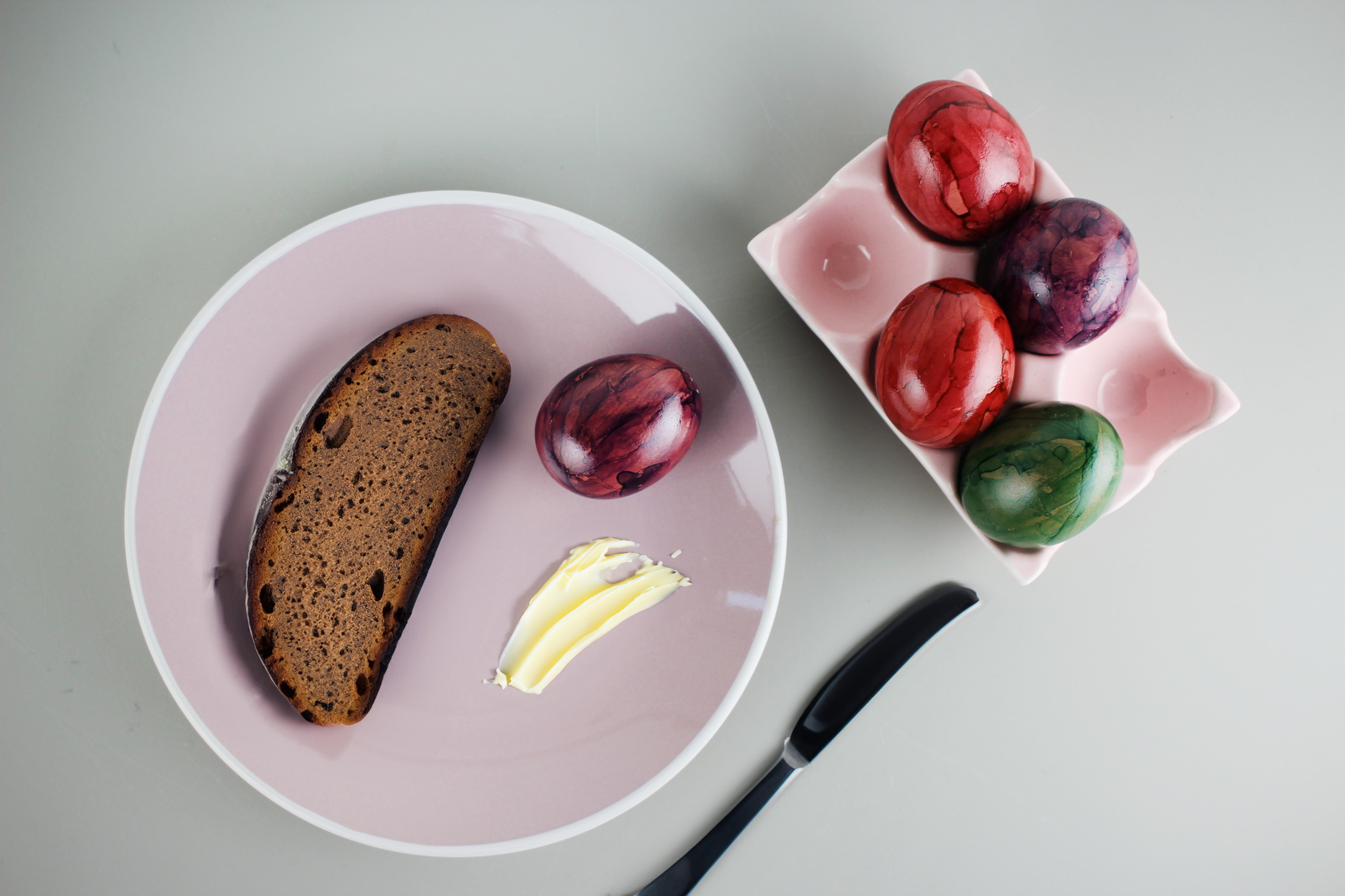 Ein Teller mit dunklem Brot, einem lila gefärbtem Ei und einem Stück Butter neben einer rosa Schale mit vier bunten Eiern.