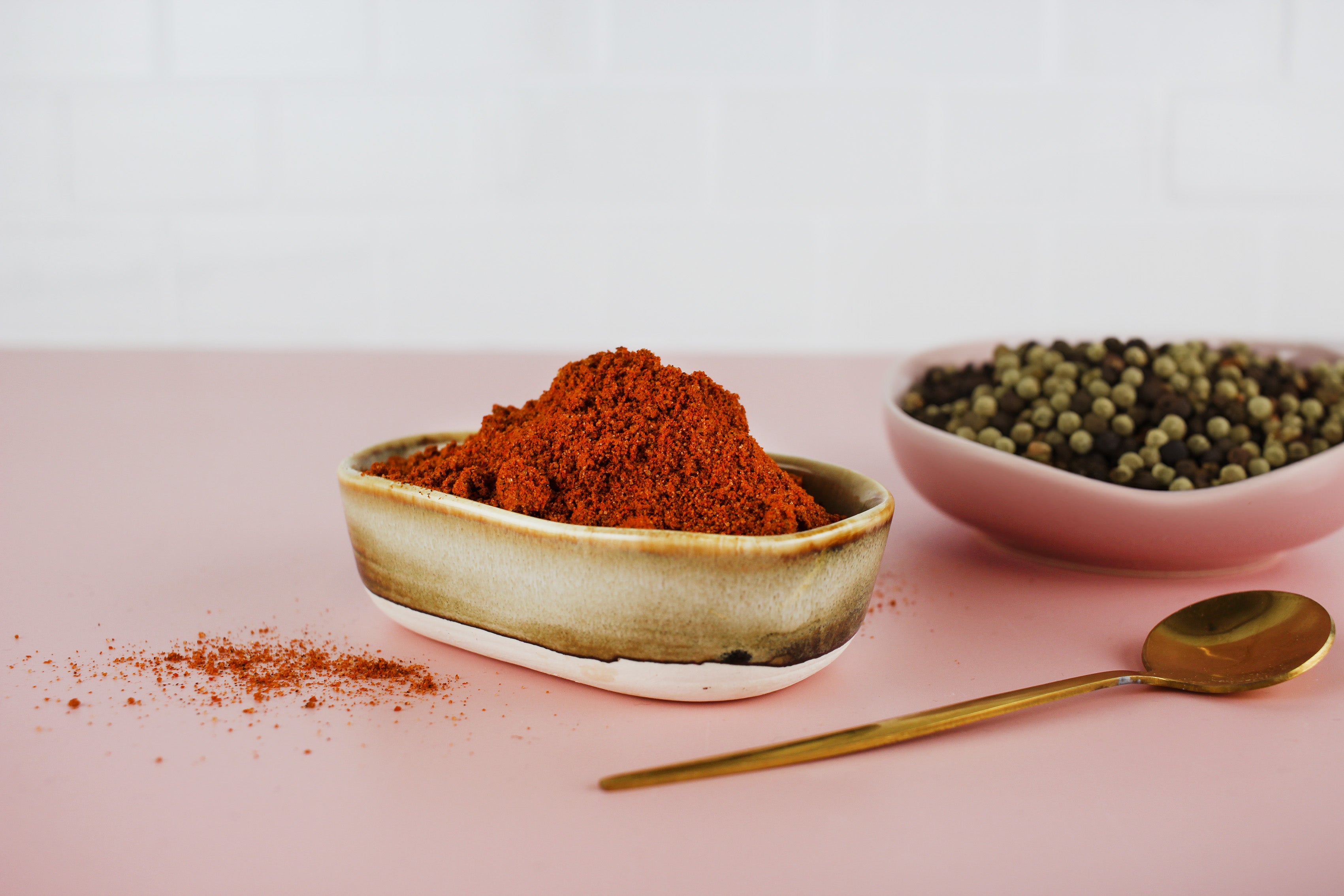 Schale mit rotem Brathähnchen-Gewürzpulver, daneben Schale mit grünen und schwarzen Pfefferkörnern und goldener Löffel auf rosa Untergrund