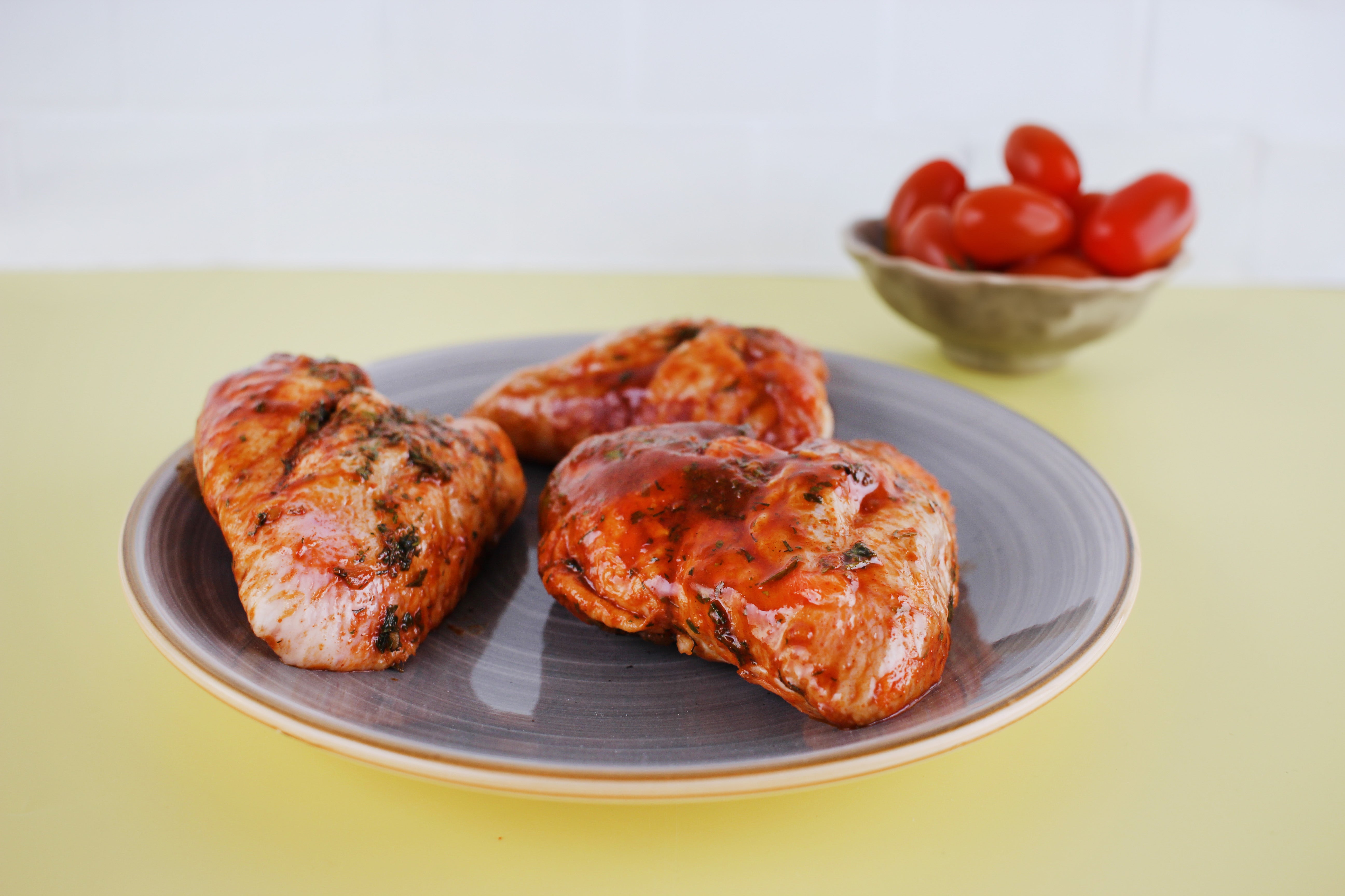 Drei gegrillte Hähnchenflügel mit glänzender Glasur liegen auf einem Teller, im Hintergrund steht eine Schale mit Kirschtomaten.