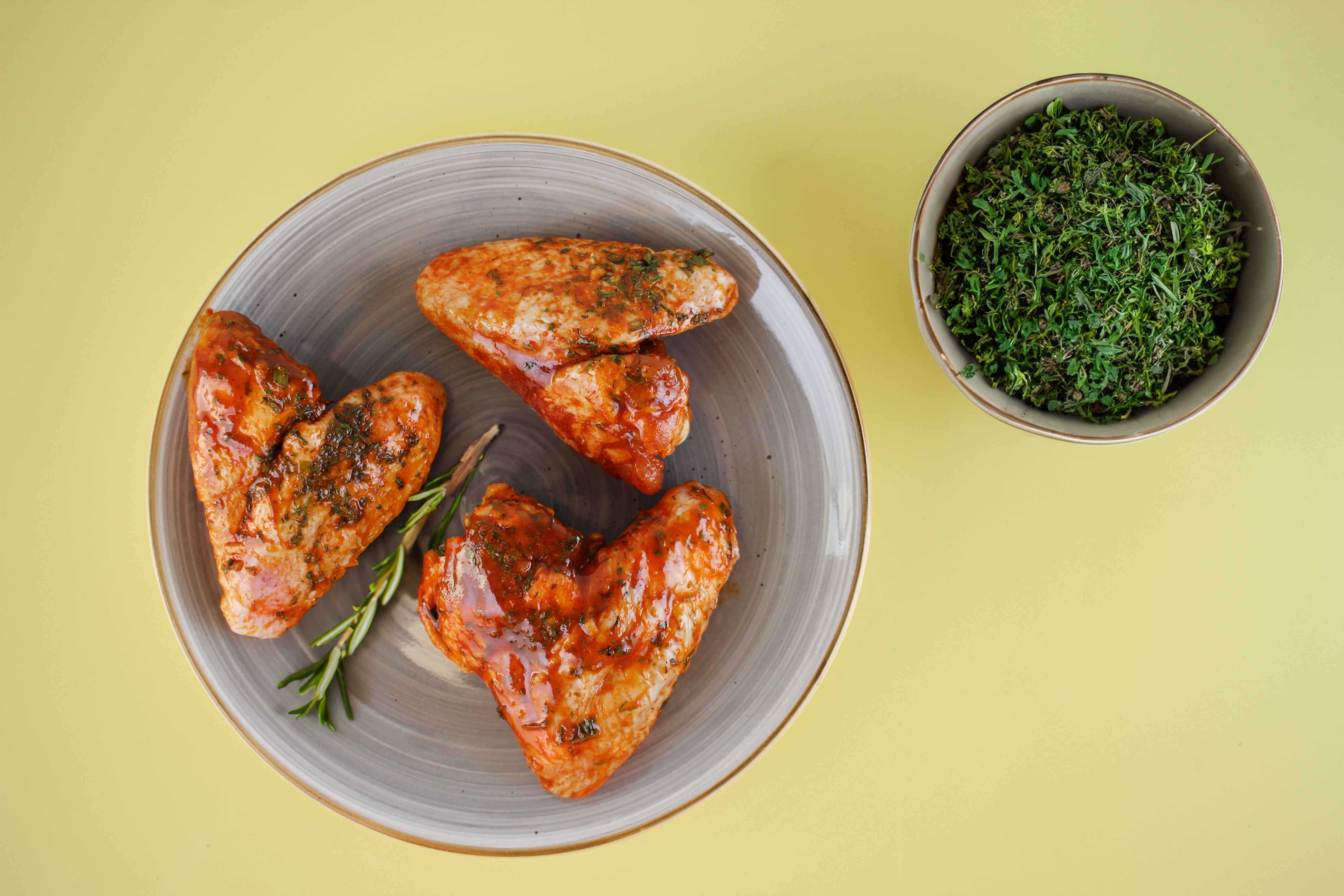 Drei glänzende, mit Sauce überzogene Hähnchenflügel auf einem Teller, dazu eine kleine Schale mit frischen Kräutern, vor einem gelben Hintergrund.