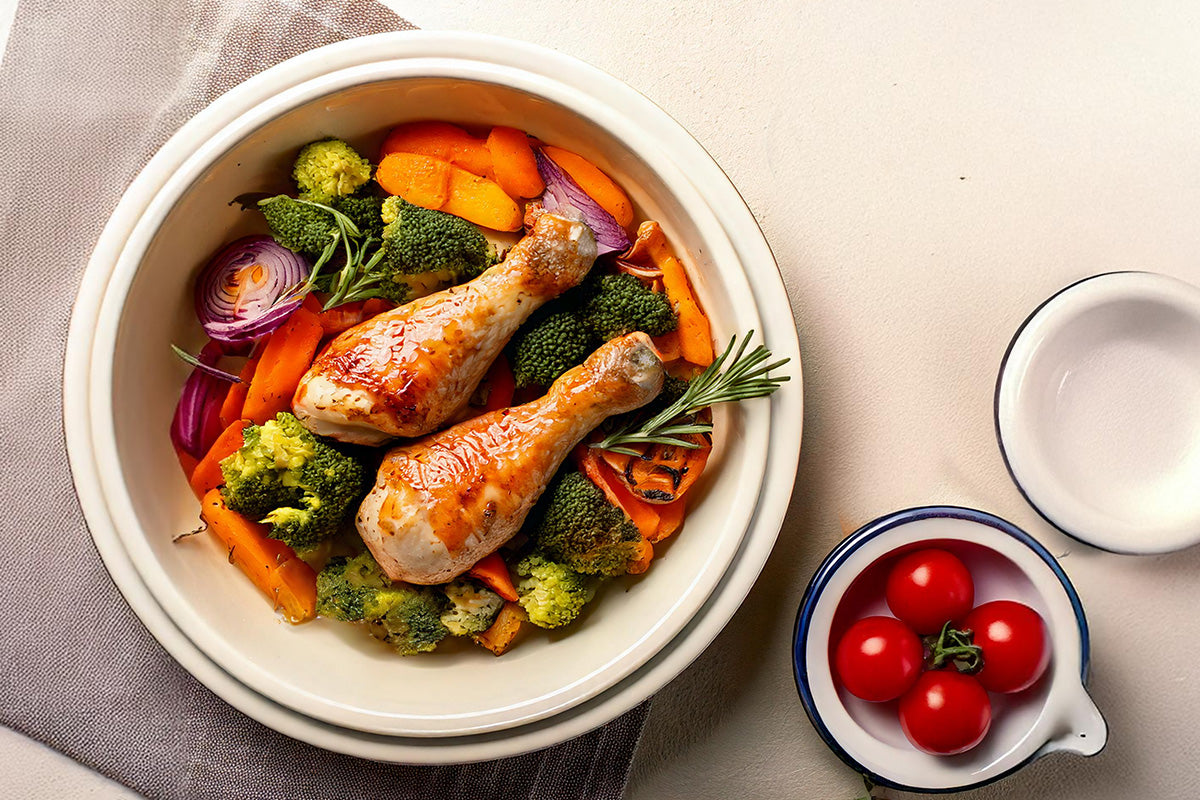 Zwei gebratene Hähnchenschenkel auf einem Bett aus Brokkoli, Karotten und roten Zwiebeln in einer weißen Schüssel, daneben eine kleine Schale mit vier Kirschtomaten