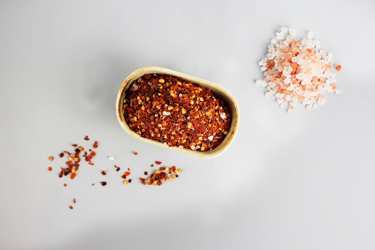 Kleiner ovaler Behälter mit roten Chiliflocken, daneben grobes rosa und weißes Salz auf hellem Untergrund.