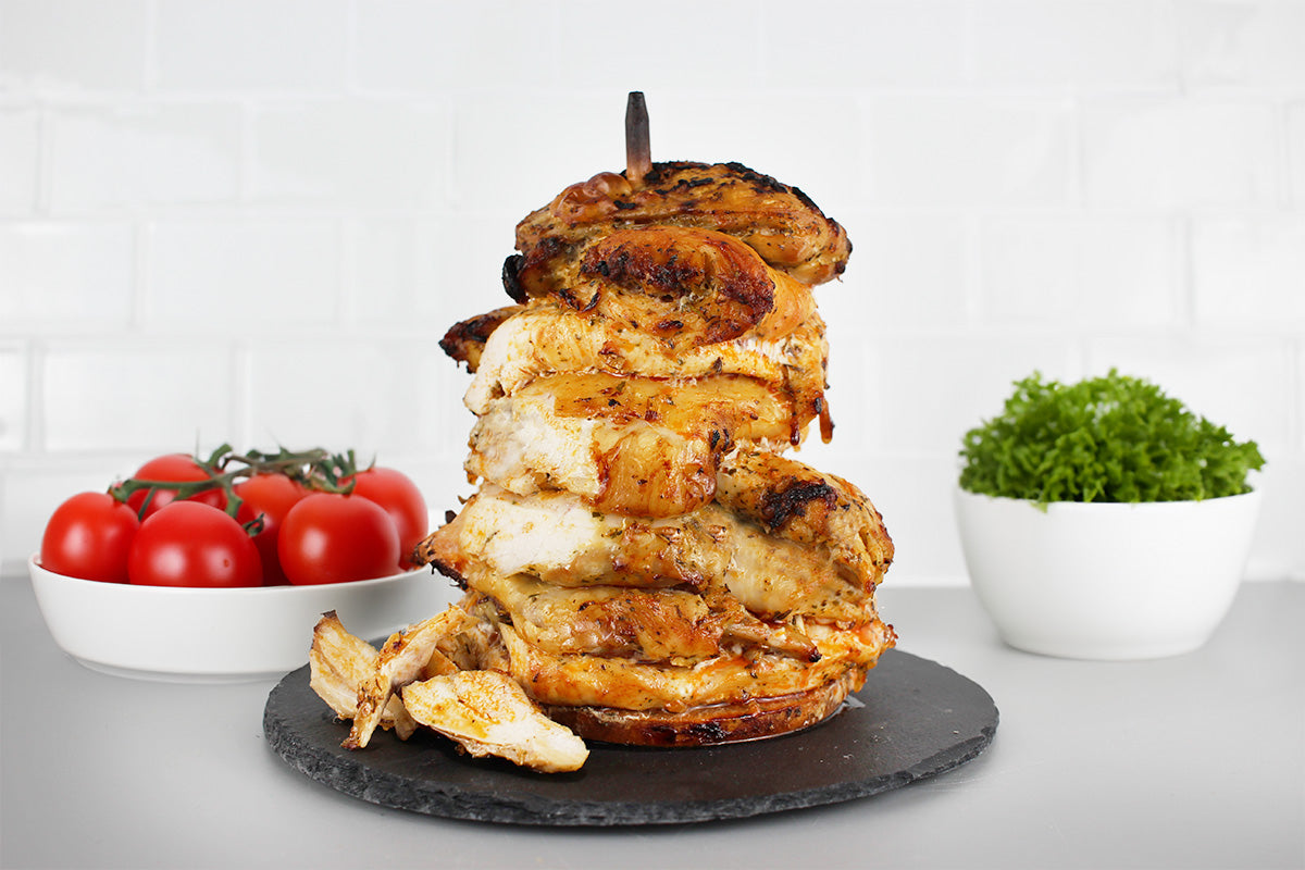 Gegrillter Hähnchendöner auf Spieß, serviert auf schwarzer Platte, im Hintergrund Schale mit Tomaten und Schale mit grünem Salat