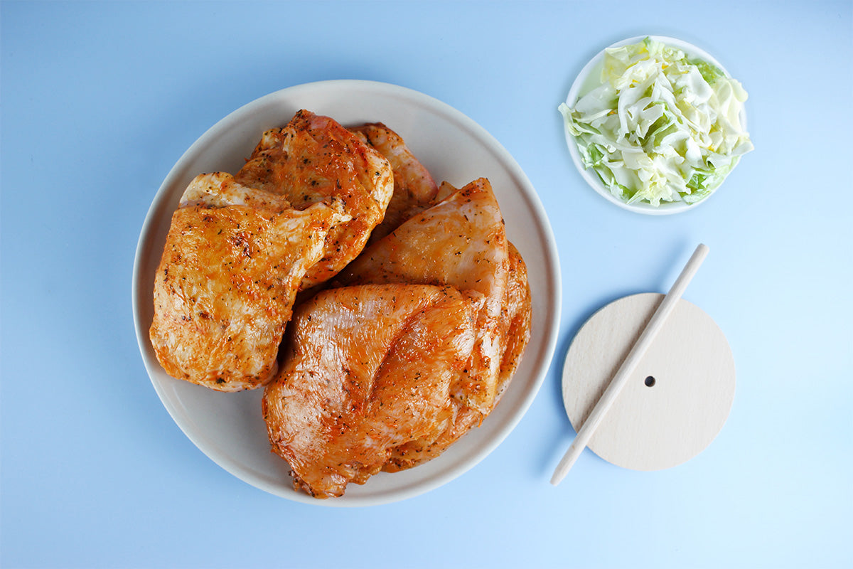 Marinierte Hähnchenfleischstücke in einer weißen Schüssel, daneben geschnittener Weißkohl in einer kleinen Schüssel und ein Dönerspieß mit Stab auf blauem Hintergrund