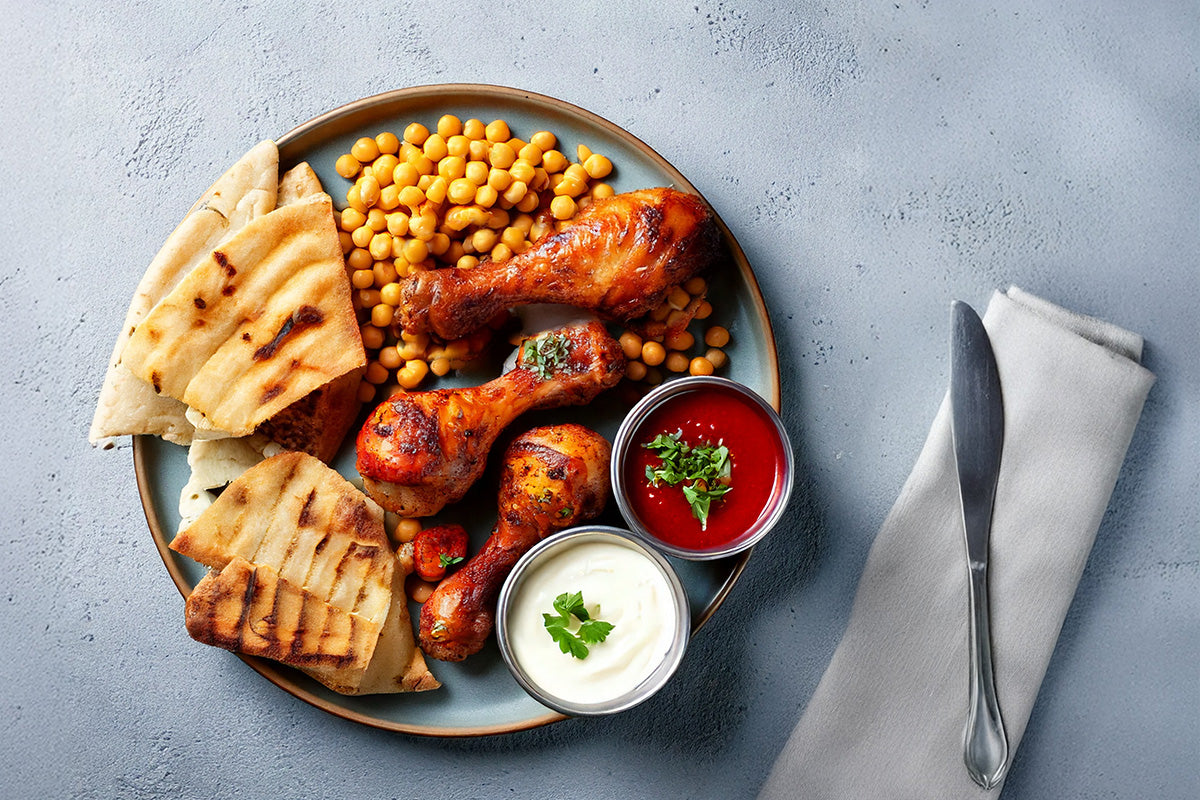 Gegrillte Hähnchenschenkel mit Paprikamarinade, Kichererbsen, Fladenbrot, rote und weiße Dips auf einem Teller, Messer auf grauem Stoff serviert