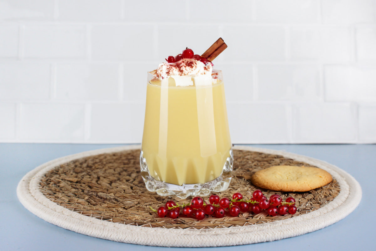 Glas mit Eierlikör, garniert mit Schlagsahne, roten Johannisbeeren und Zimtstange, daneben ein Keks und rote Johannisbeeren auf einem runden Untersetzer