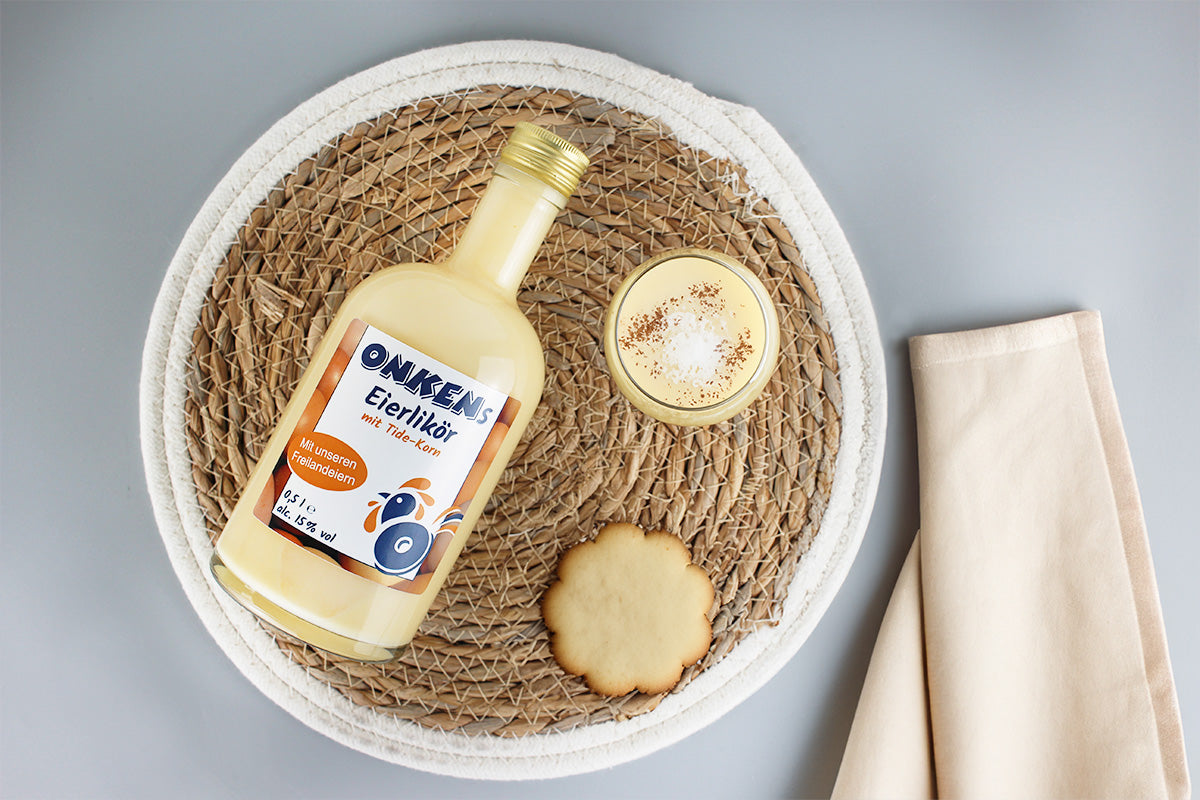 Flasche Onkens Eierlikör mit Tide-Korn und Freilandeiern, Glas Eierlikör mit Gewürz und Keks auf rundem geflochtenem Untersetzer, beige Serviette daneben