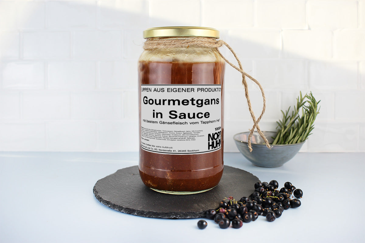Glas mit Gourmetgans in Sauce, beschriftet mit 'Gourmetgans in Sauce', daneben schwarze Johannisbeeren und ein Schälchen mit Rosmarin