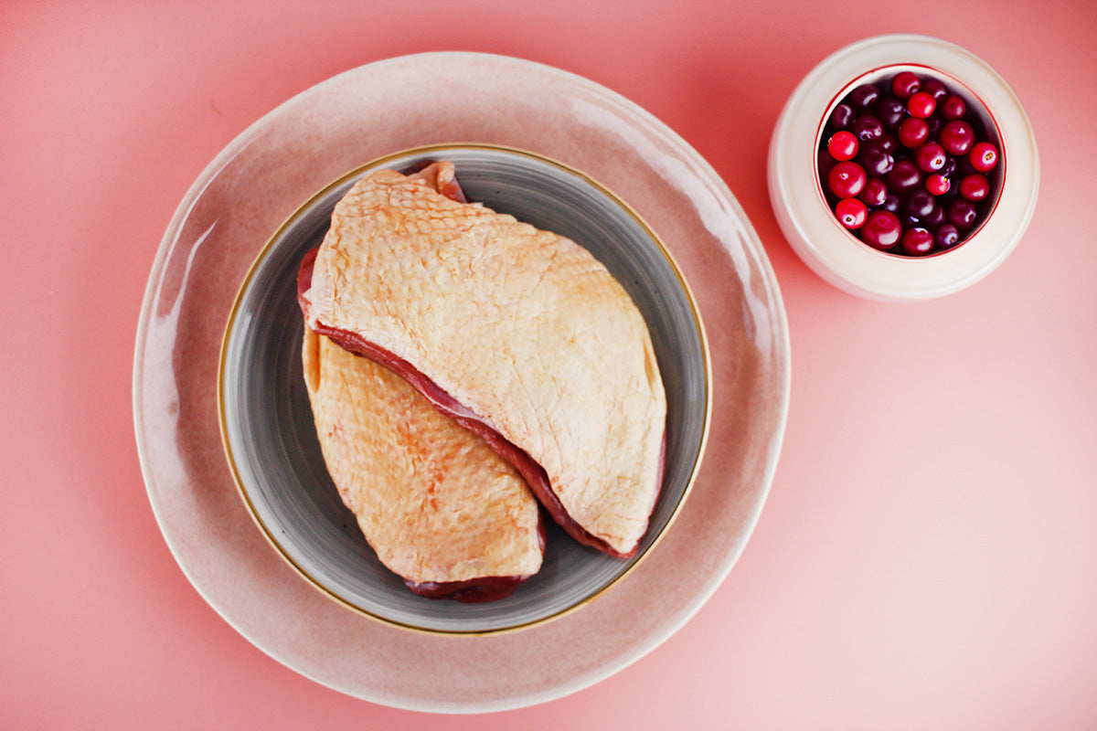 Zwei rohe Entenbrustfilets mit Haut auf einem grauen Teller, daneben eine Schale mit frischen roten Preiselbeeren auf rosa Untergrund