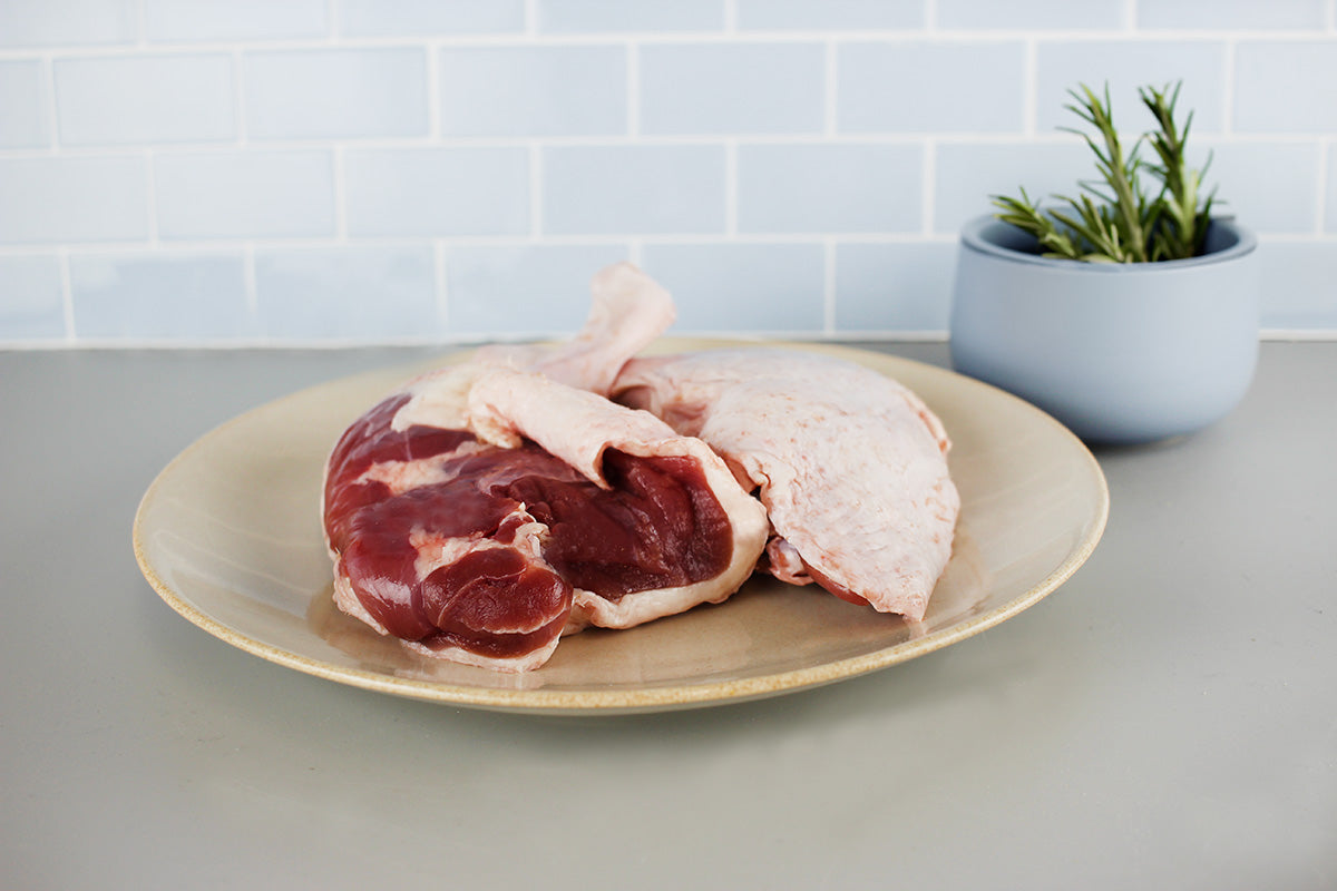 Rohes Entenkeulenfleisch auf einem hellen Teller, im Hintergrund ein Topf mit frischen Kräutern