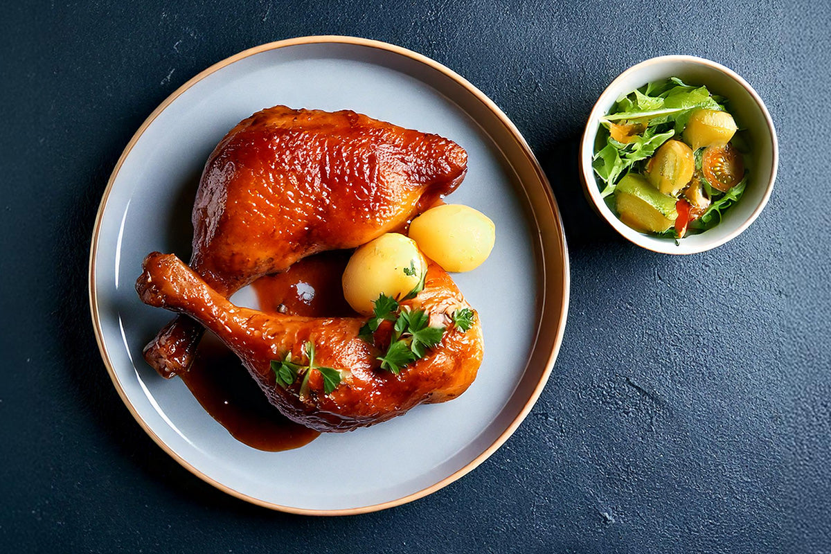 Zwei gebratene Entenkeulen mit Bratensauce und zwei Kartoffeln auf einem weißen Teller, daneben eine kleine Schüssel mit grünem Salat und Gurkenscheiben