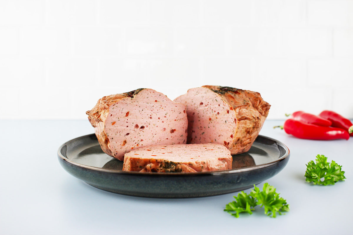 Aufgeschnittener Fleischkäse mit Kräutern auf dunklem Teller, daneben rote Chilischoten und grüne Petersilienzweige.