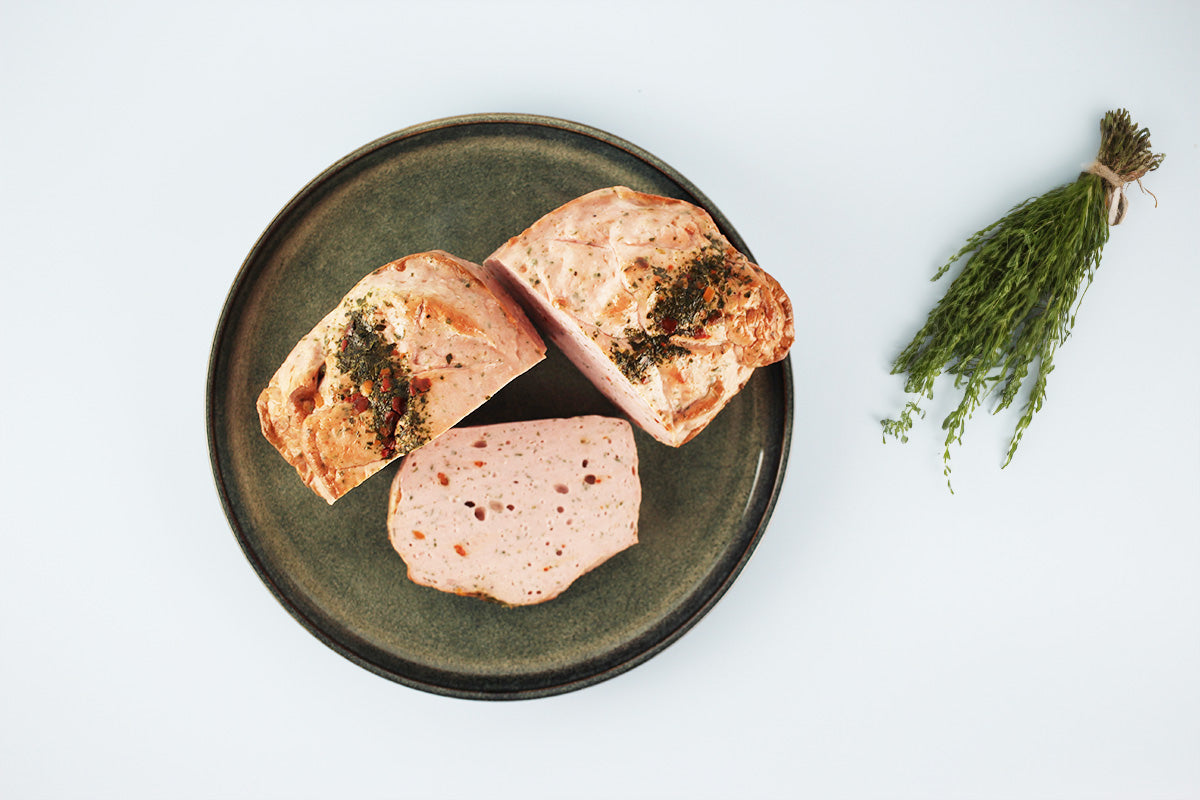 Drei Stücke Fleischkäse auf einem dunklen Teller, daneben ein Bündel frischer Kräuter mit Bindfaden.