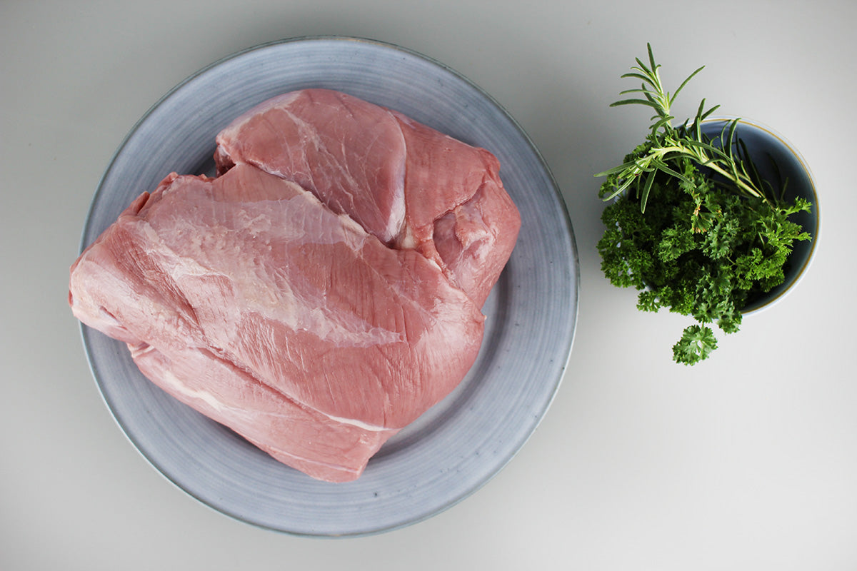 Rohe Frischlingskeule auf grauem Teller neben Schale mit frischem Rosmarin und Petersilie