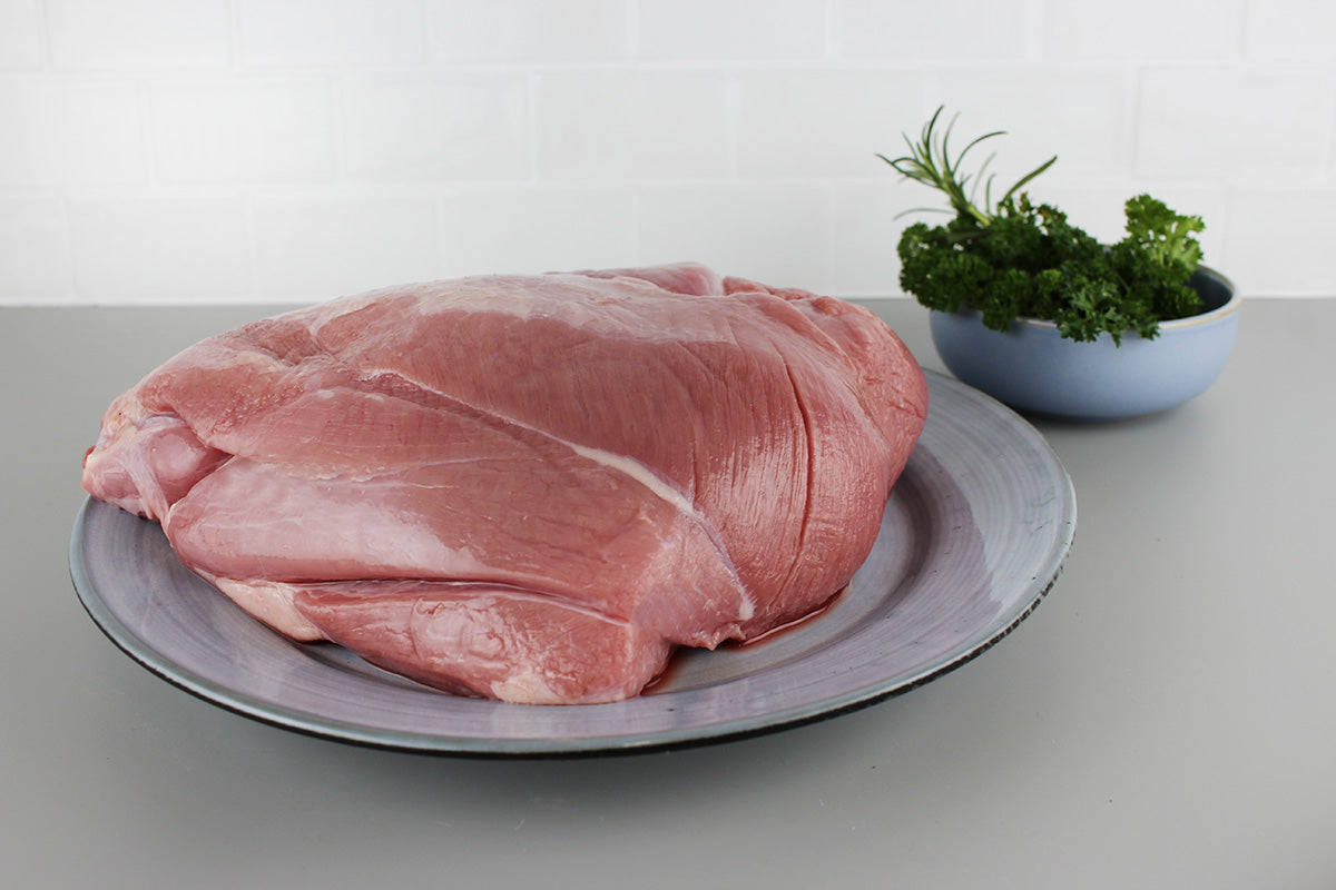 Rohe Frischlingskeule auf grauem Teller, im Hintergrund Schale mit frischen Kräutern