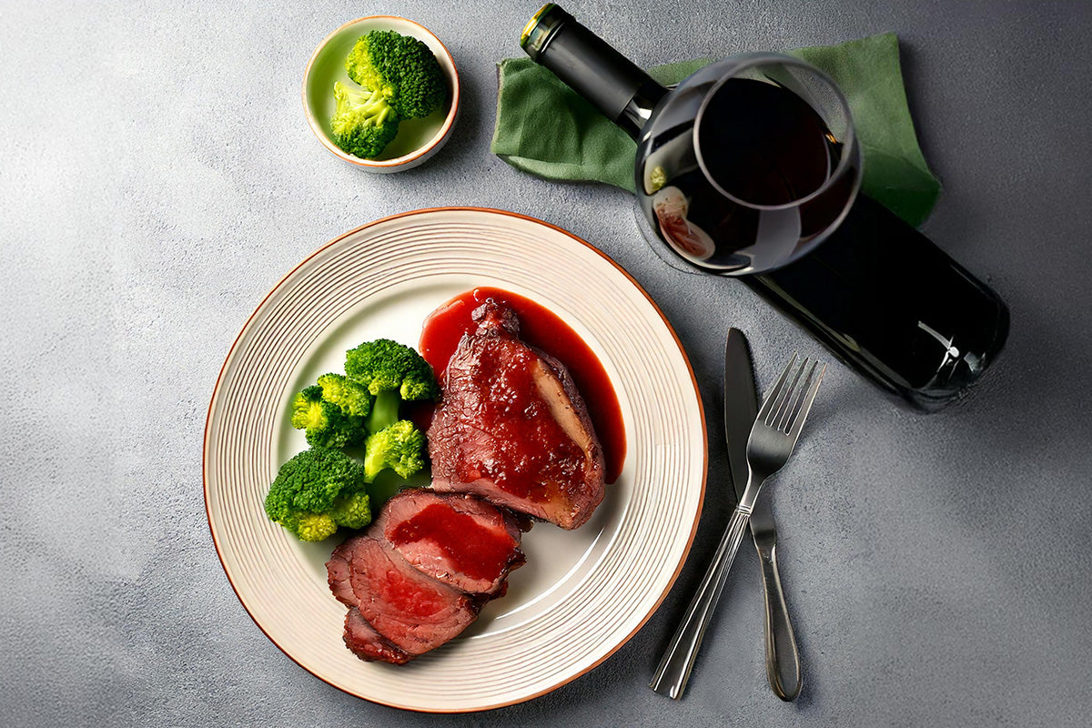 Frischlingskeule mit roter Sauce und gedämpftem Brokkoli auf weißem Teller, daneben Flasche und Glas Rotwein, Messer und Gabel