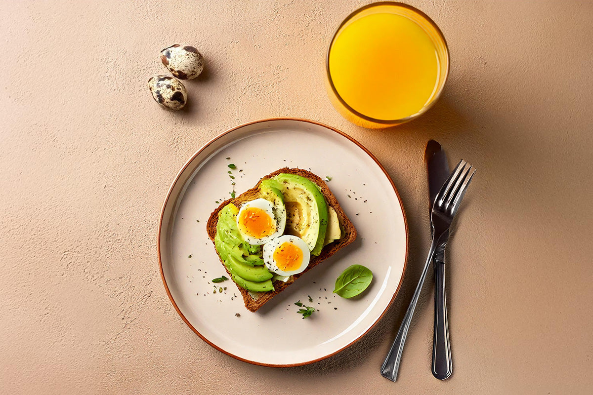 Vollkornbrot mit Avocadoscheiben und halbierten Wachteleiern auf weißem Teller, daneben zwei Wachteleier, Glas Orangensaft, Messer und Gabel