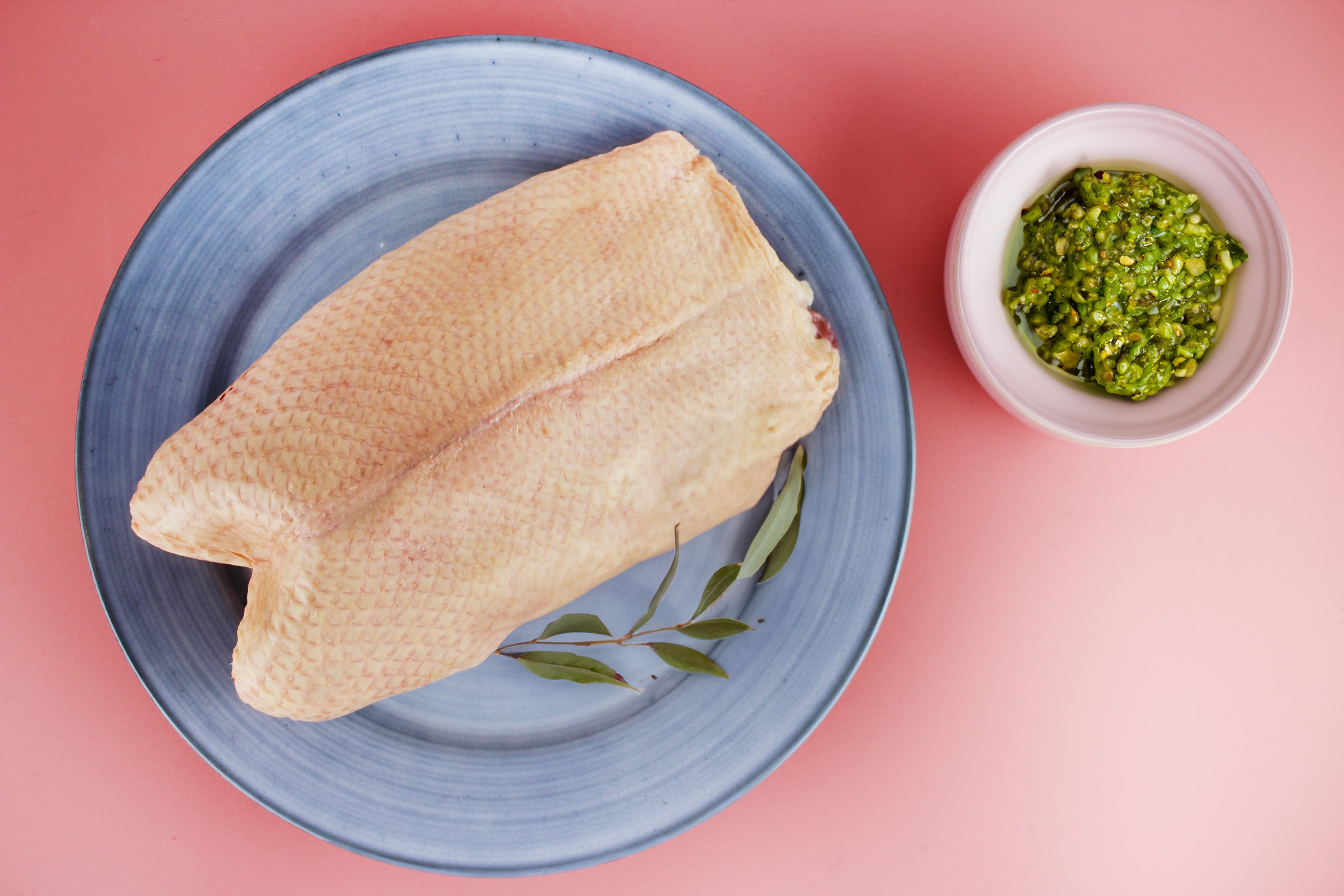 Rohe Gänsebrust auf blauem Teller mit Lorbeerblättern, daneben kleine Schüssel mit grüner Kräuterpaste auf rosa Untergrund