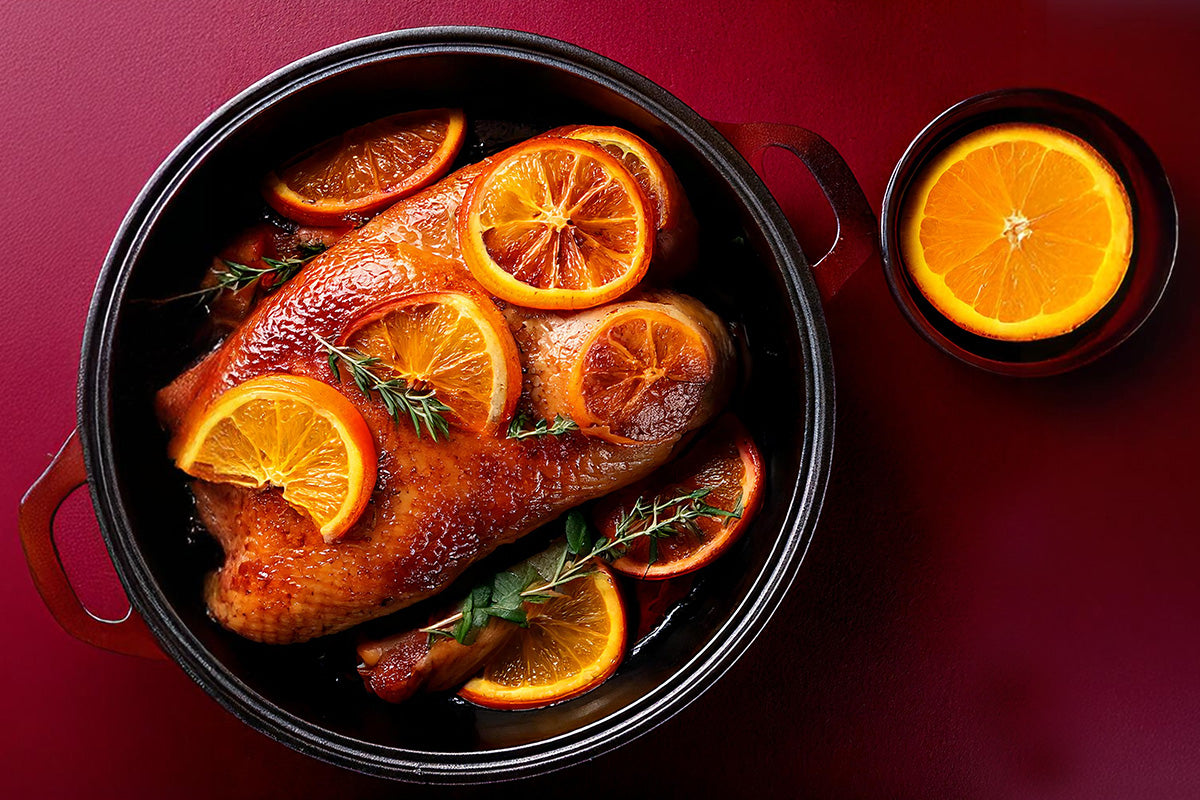 Gänsebrust im Bräter mit Orangenscheiben und Kräutern in Orangensauce, daneben eine Schale mit einer Orangenscheibe