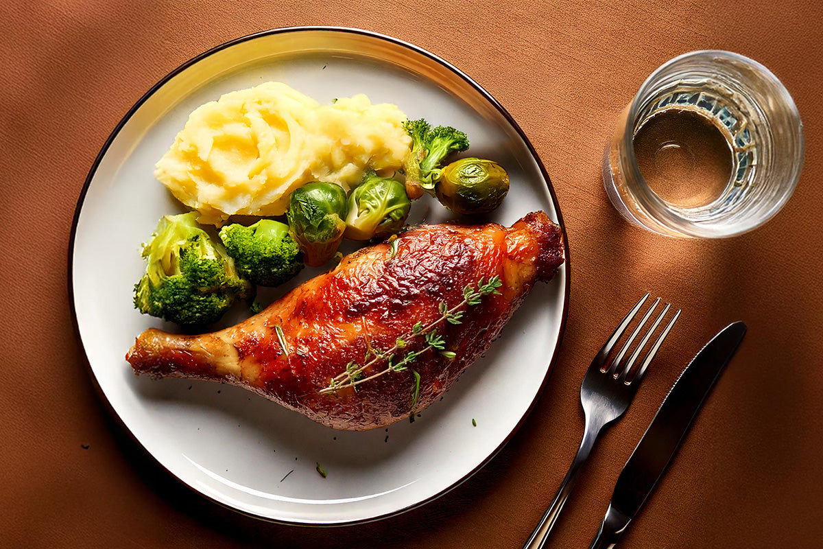 Gebratene Gänsebrust mit Kartoffelpüree, Rosenkohl und Brokkoli auf weißem Teller, daneben Glas Wasser, Messer und Gabel