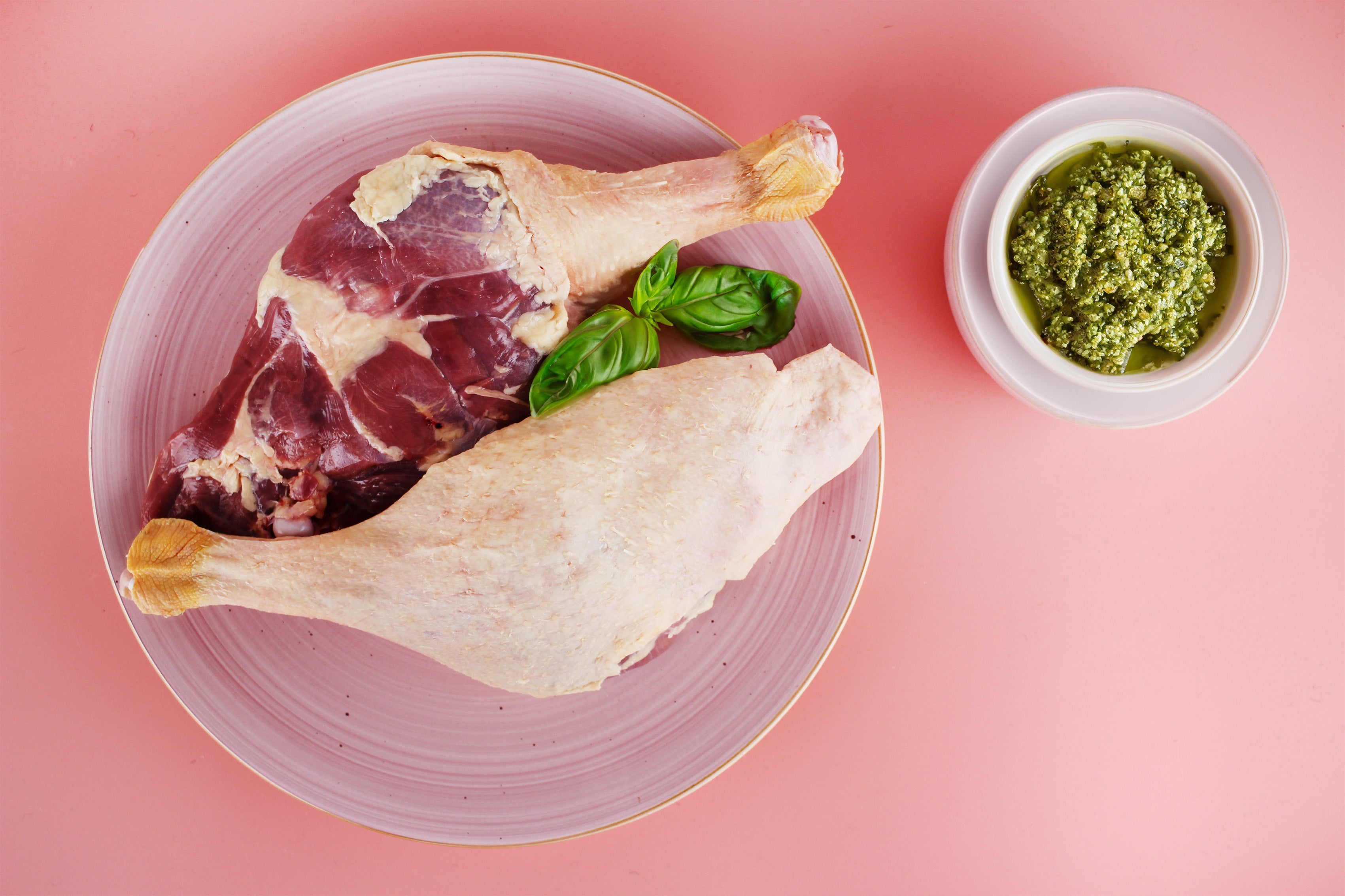 Zwei rohe Gänsekeulen auf einem Teller mit Basilikumblättern, daneben eine Schale mit grünem Pesto auf rosa Untergrund