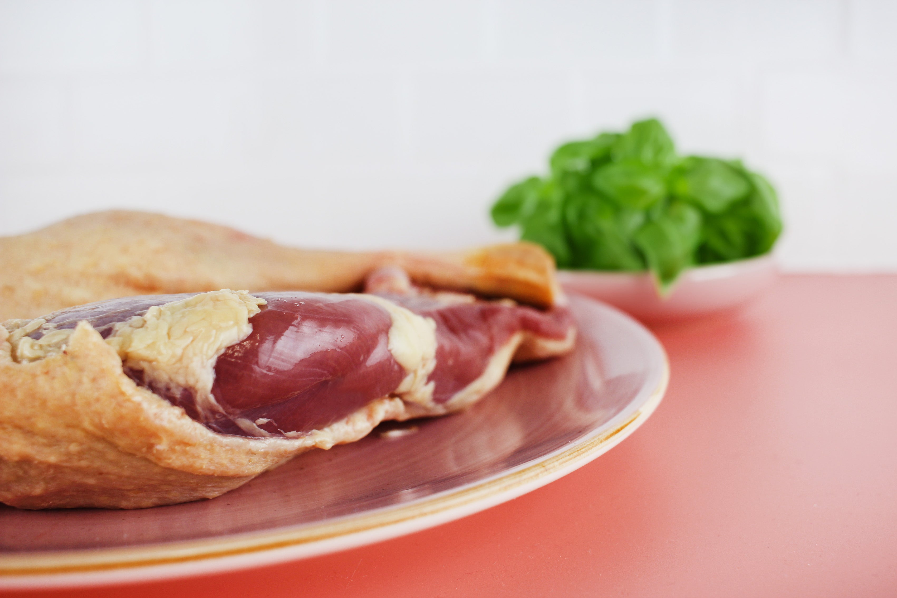 Rohe Gänsekeule mit Haut auf einem Teller, im Hintergrund Schale mit frischem grünem Basilikum