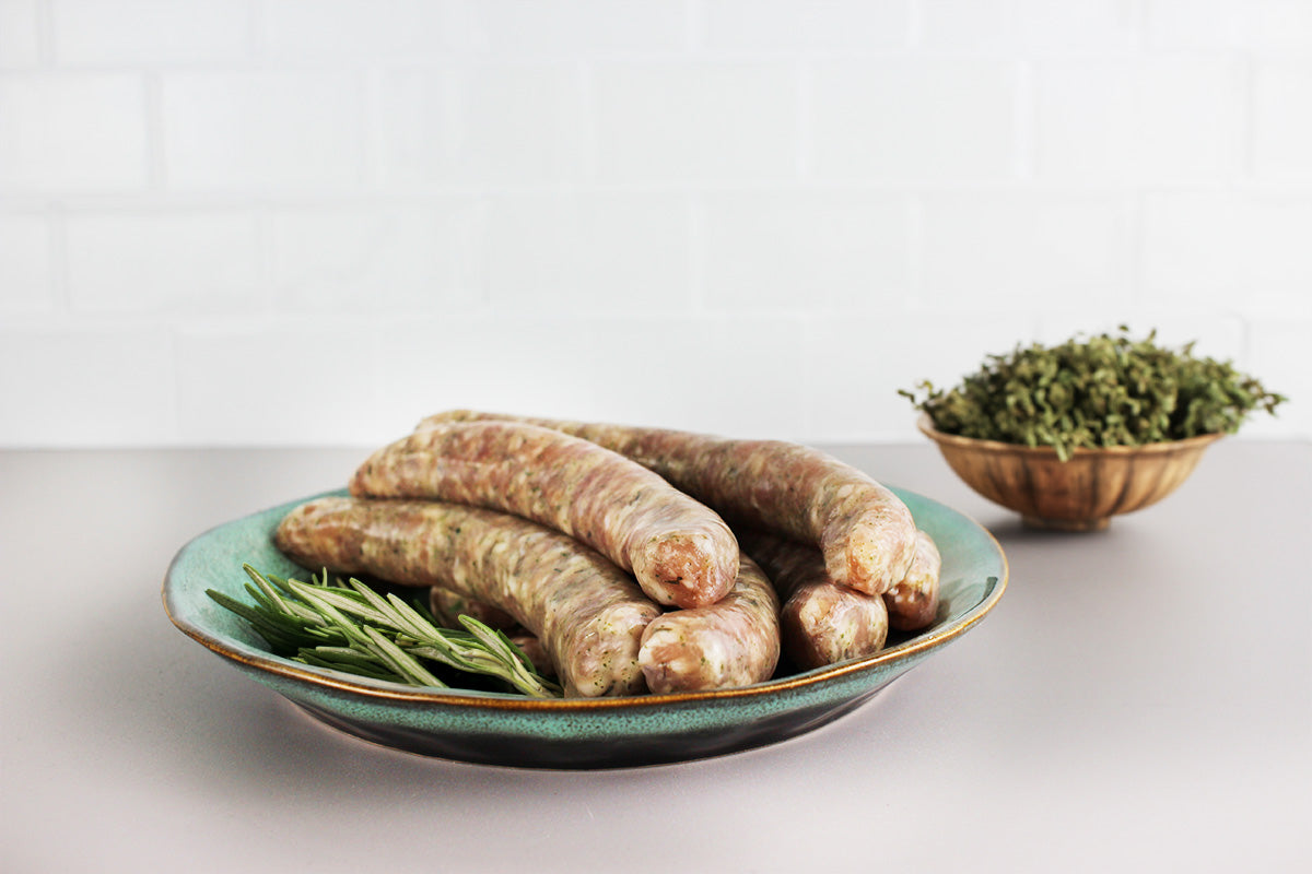 Sechs rohe Geflügel-Chipolata-Würste auf einem grünen Teller mit Rosmarinzweigen, im Hintergrund eine Schale mit getrockneten Kräutern