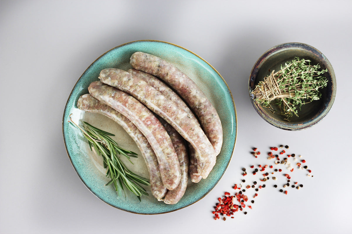 Fünf rohe Geflügel-Chipolata-Grillwürste auf einem blauen Teller mit Rosmarin, daneben ein Bund Thymian in einer Schale und bunte Pfefferkörner auf weißem Untergrund