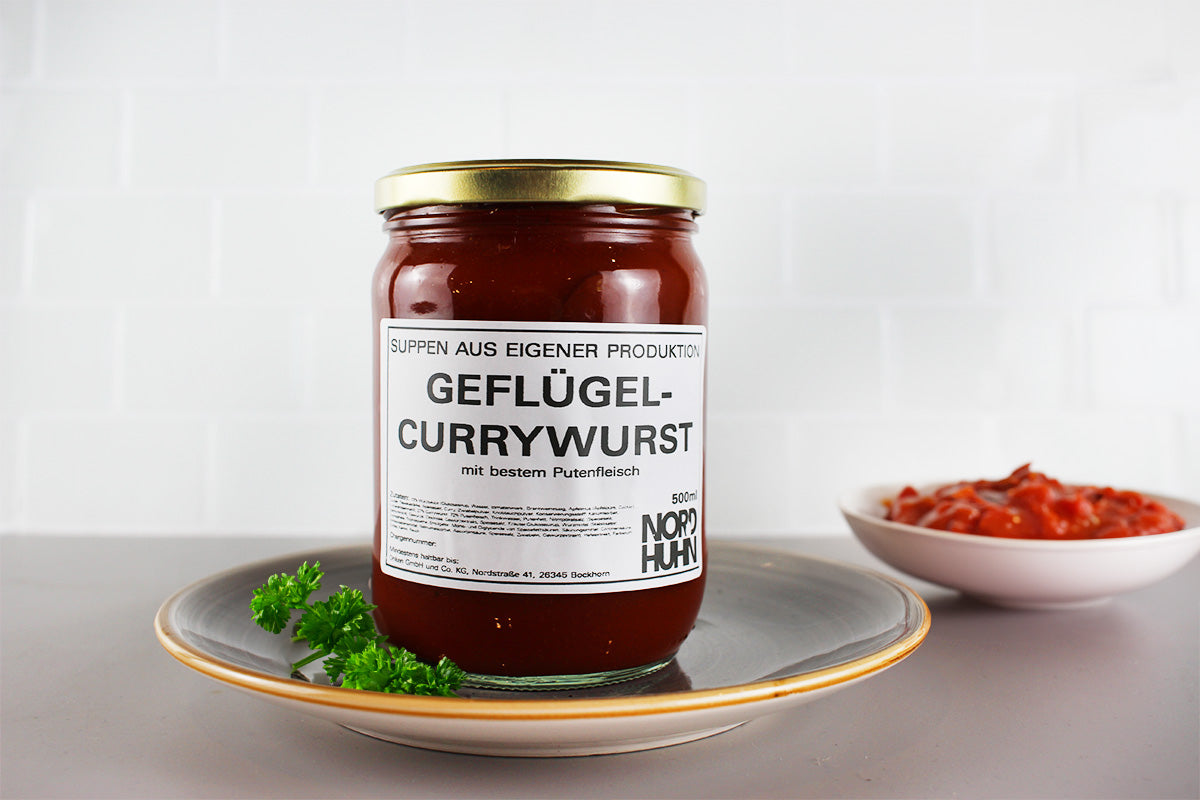 Ein Glas mit Geflügel-Currywurst in roter Sauce steht auf einem grauen Teller mit einem Stück Petersilie daneben, im Hintergrund eine Schale mit roter Sauce