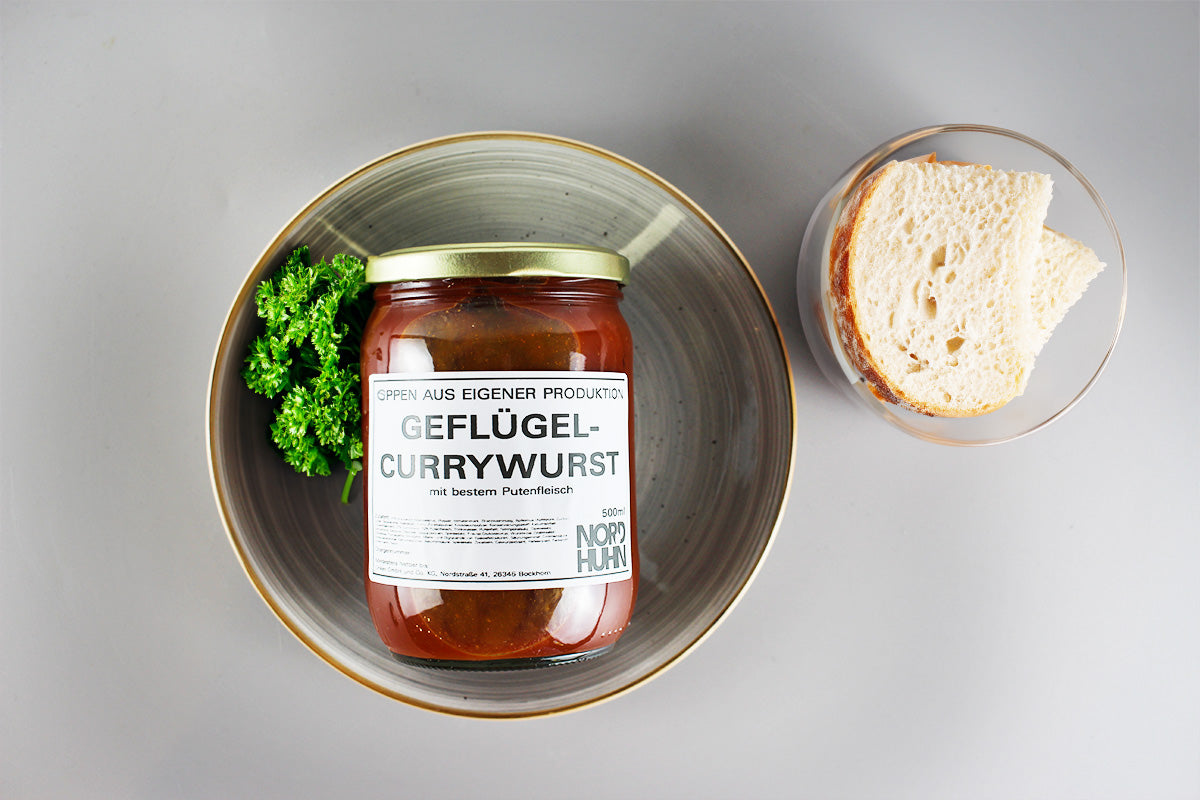 Ein Glas Geflügel-Currywurst mit Etikett auf einem grauen Teller neben einem Stück Brot und etwas Petersilie