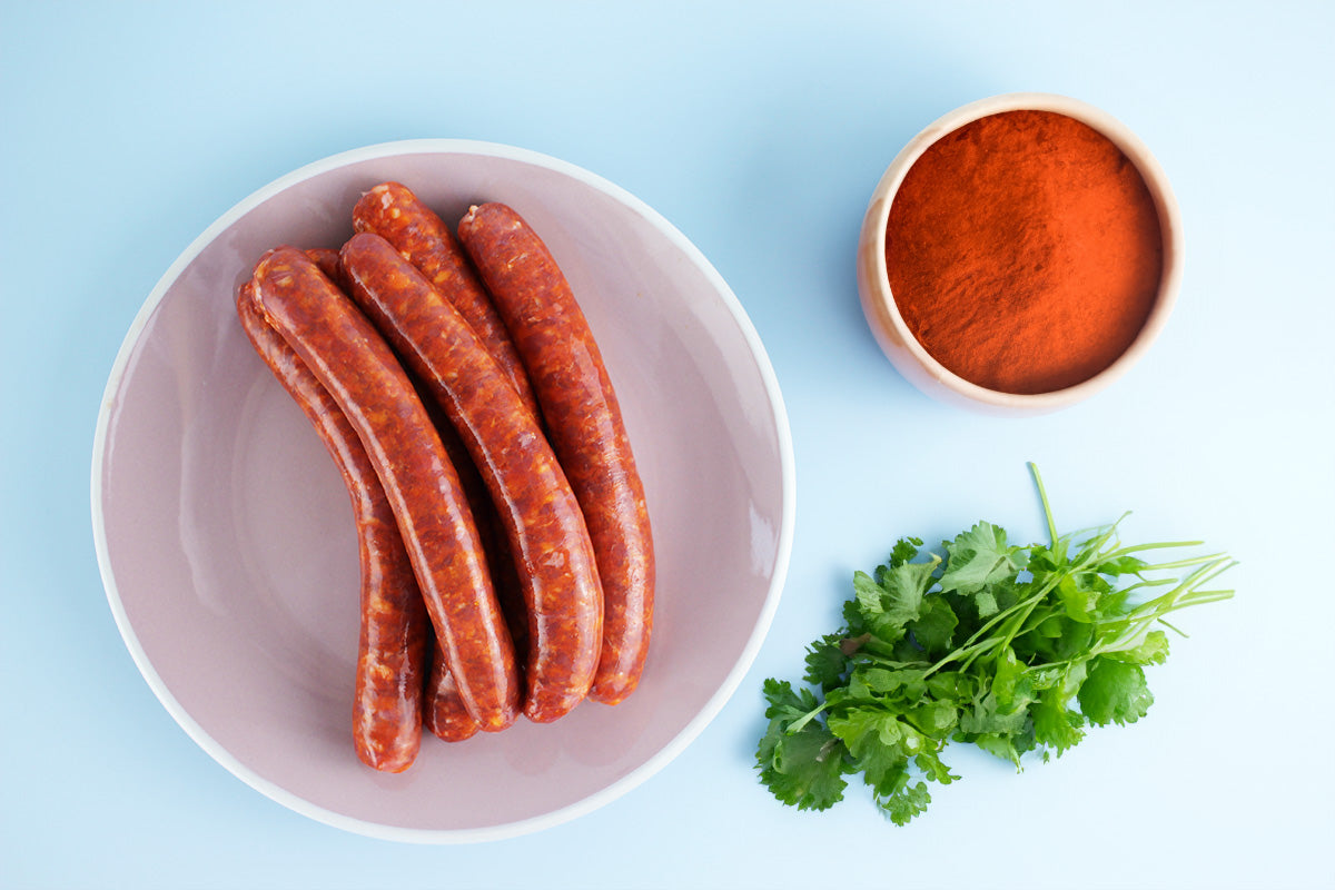 Sechs rote Geflügel-Merguez-Würste auf einem rosa Teller, daneben eine Schale mit rotem Paprikapulver und frischer Koriander auf hellblauem Hintergrund