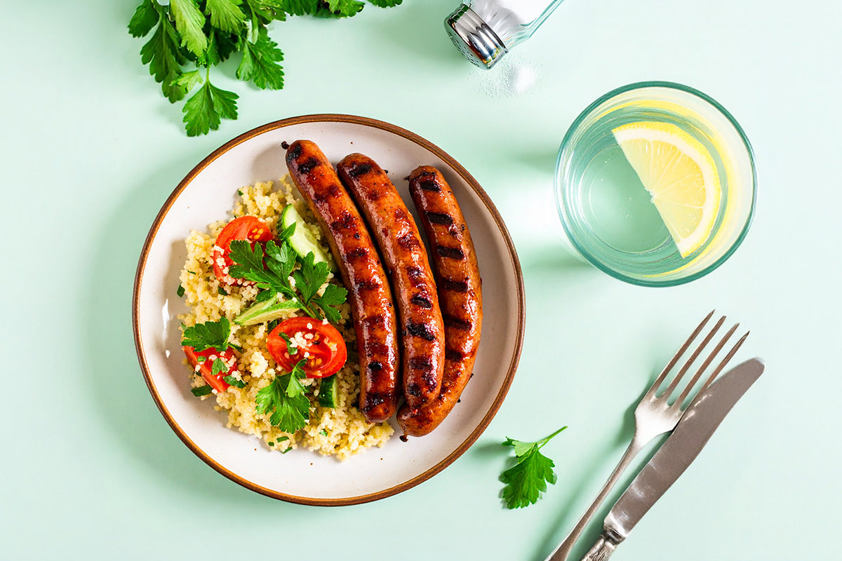 Drei gegrillte Merguez-Würste auf einem Teller mit Couscous, Tomaten, Gurken und Petersilie, daneben ein Glas Wasser mit Zitronenscheibe und Besteck
