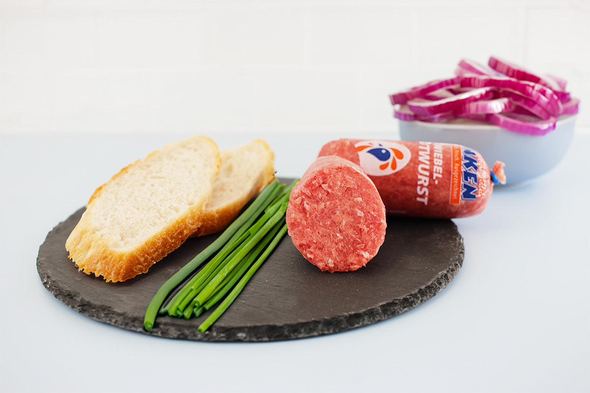 Geflügel Zwiebelmettwurst auf schwarzer Schieferplatte mit Schnittlauch, zwei Scheiben Brot und Schüssel mit roten Zwiebelringen