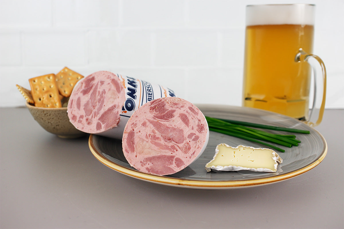 Aufgeschnittener Geflügel-Bierschinken auf einem grauen Teller mit Schnittlauch, einem Stück Brie-Käse, einer Schale mit Crackern und einem Bierglas im Hintergrund