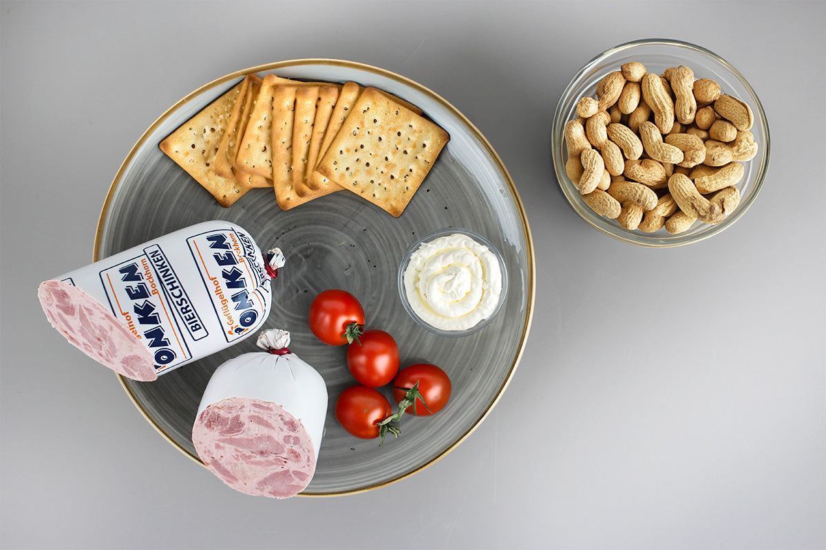 Grauer Teller mit Geflügelbierschinken, Crackern, Frischkäse und Kirschtomaten, daneben Schüssel mit Erdnüssen in Schale