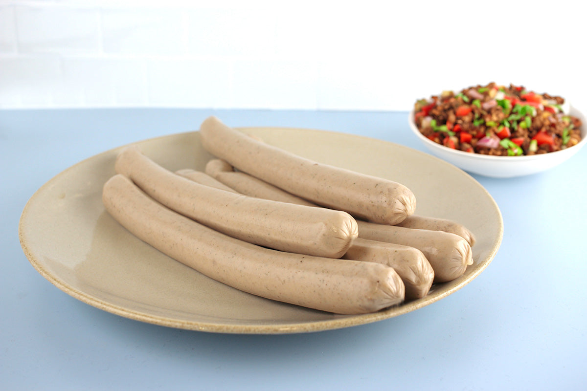 Sechs ungekochte helle Geflügelbratwürste auf einem großen weißen Teller, im Hintergrund eine Schüssel mit gehacktem Gemüse und Kräutern