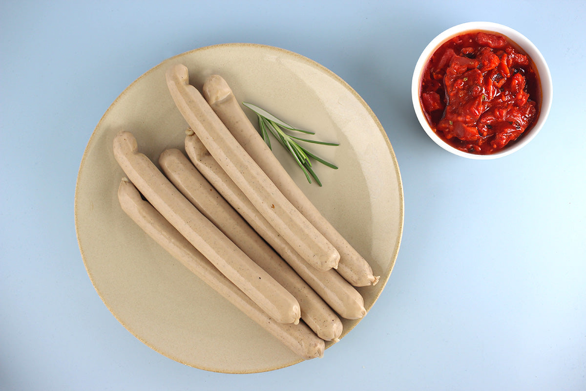 Sechs helle Geflügelgrillwürstchen auf einem beigen Teller mit Rosmarin, daneben eine Schale mit rotem Tomatensugo auf hellblauem Hintergrund