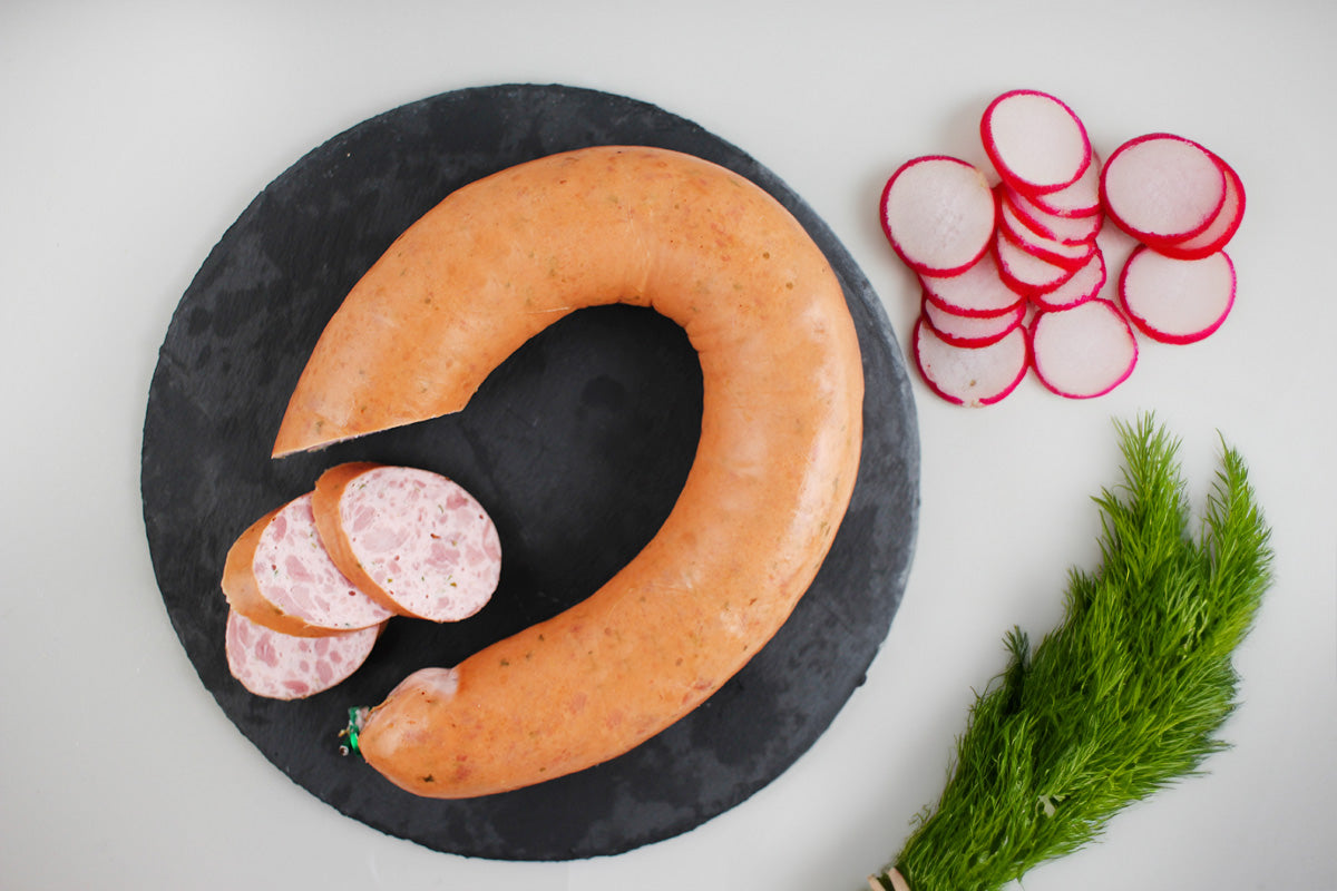 Ringförmige Geflügelwurst mit mehreren Scheiben auf schwarzem runden Untergrund neben Radieschenscheiben und Dillzweigen